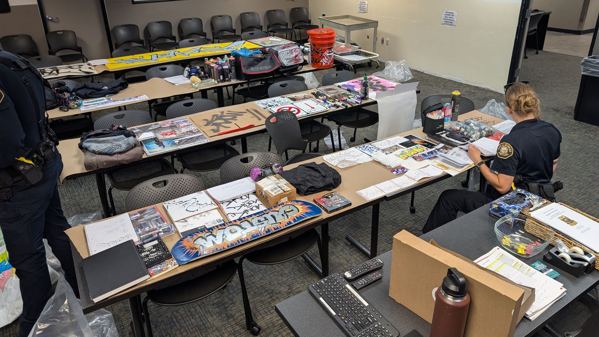 Tables covered with art supplies, graffiti sketches, spray paint cans, and skateboards in a room with police officers reviewing materials and empty chairs around.