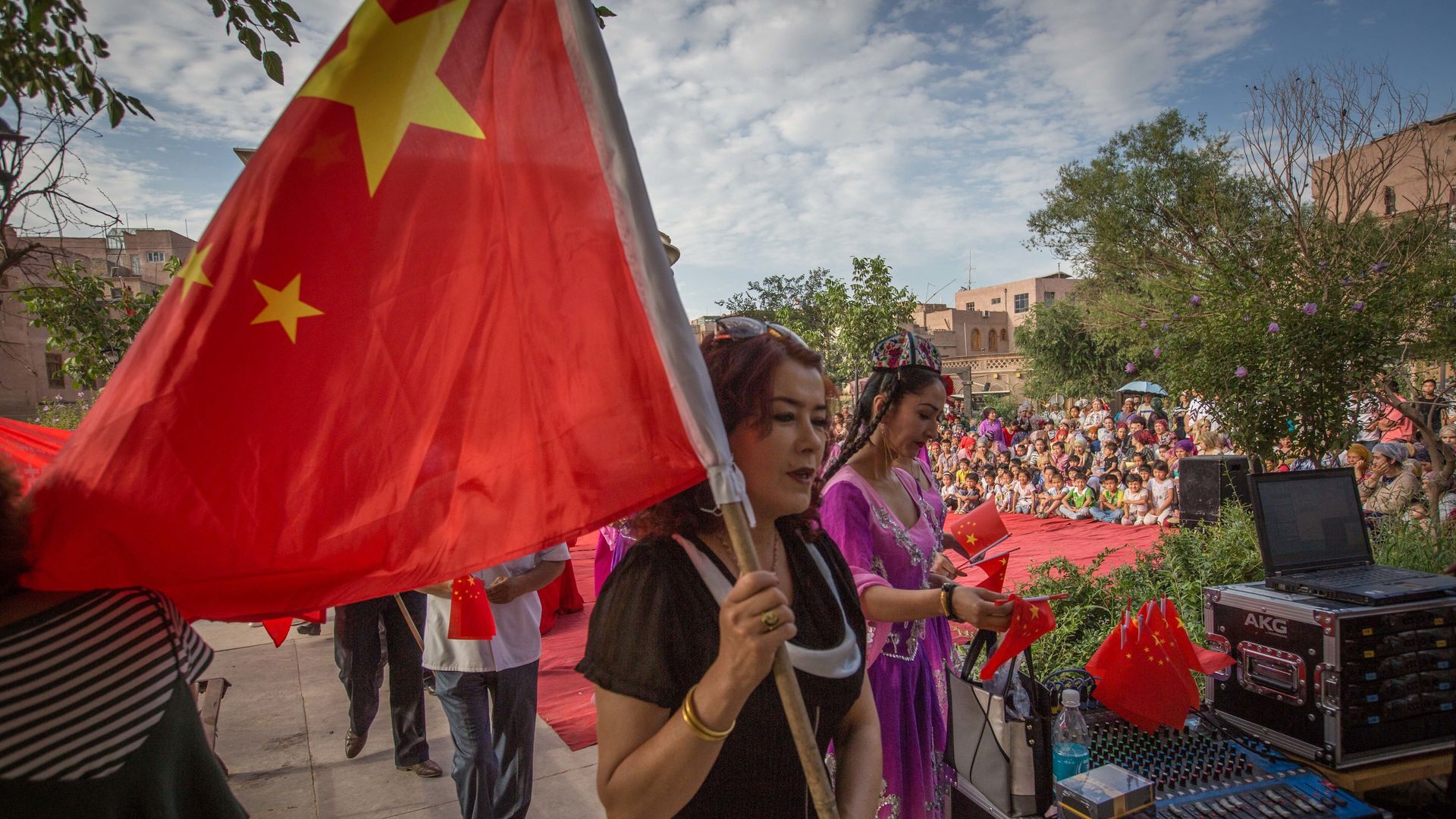 In this image, a Uyghur woman in a black shirt holds a Chinese flag above her as she walks down the street on a cloudy day.