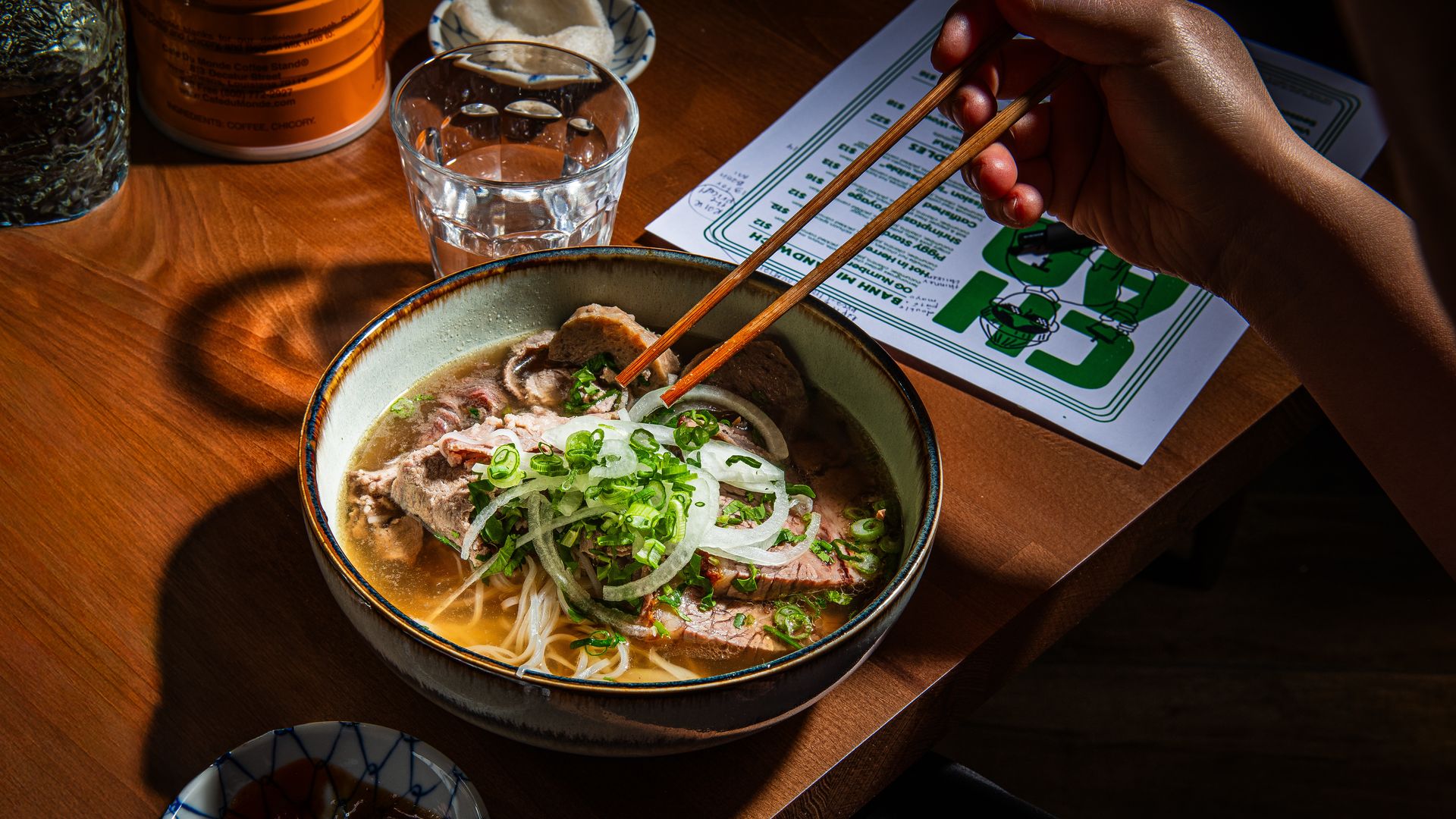 Hand using chopsticks to pick up food from a bowl of pho with sliced beef, onions, and green onions on a wooden table with a glass of water, a menu, and a small dish of sauces.