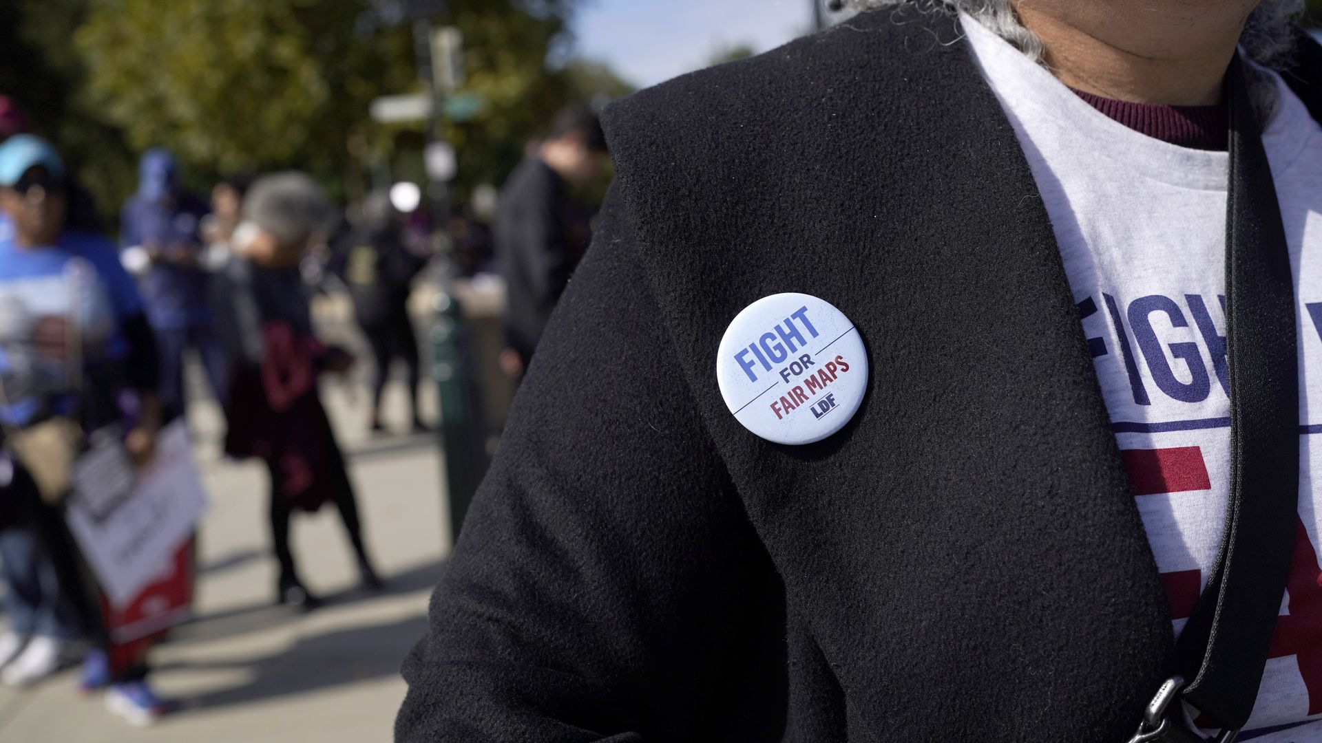 A button worn by a protester outside the Supreme Court that reads, "Fight for fair maps," with several other protesters in the background.