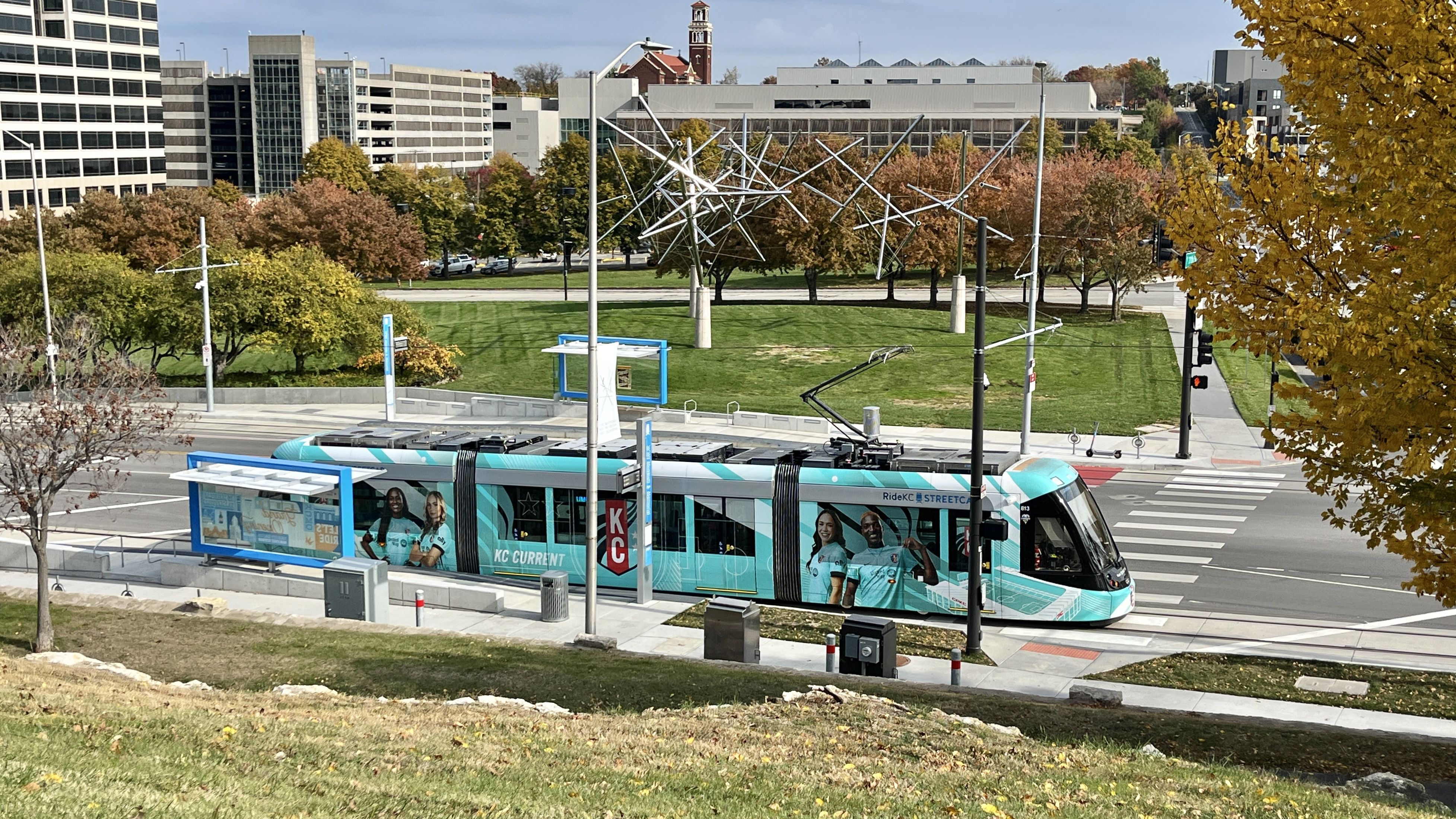 A teal KC Current streetcar at a stop in an urban area with modern buildings, green lawn, and trees with autumn foliage around. Silver abstract sculpture visible in background.