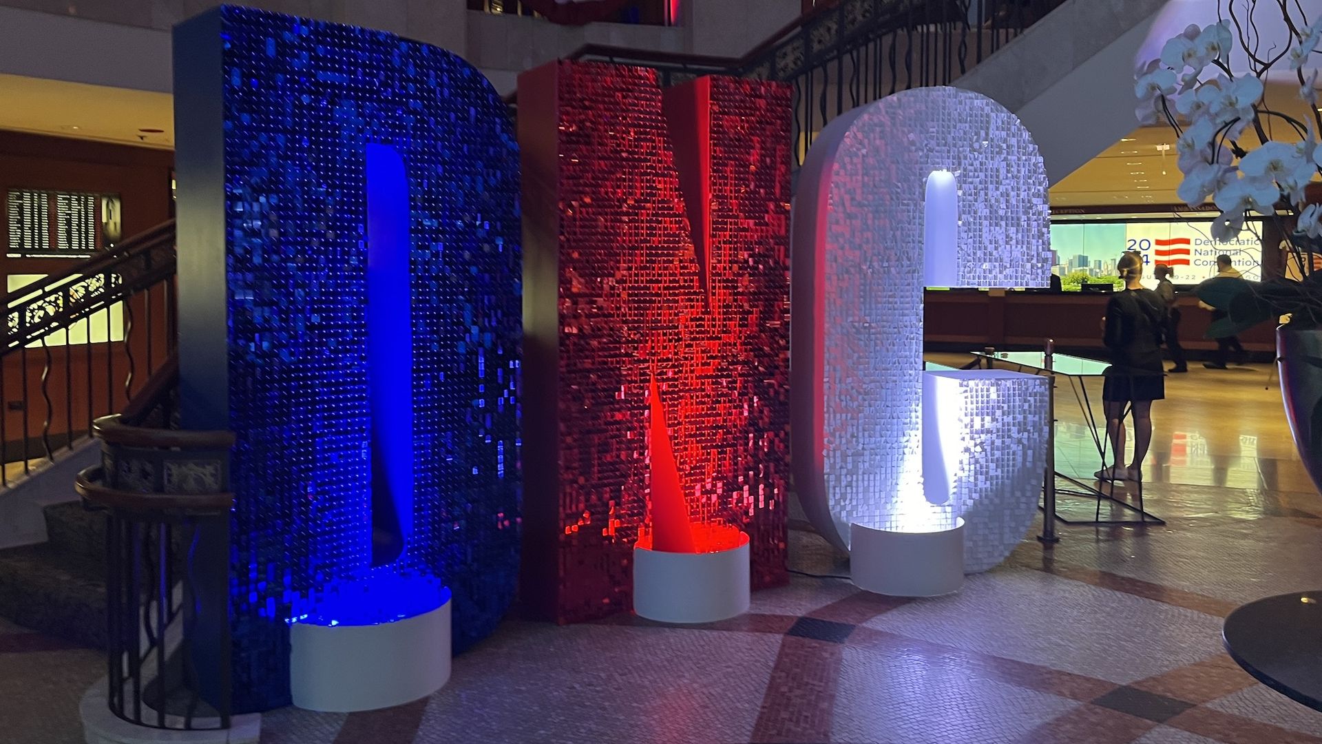 Large illuminated letters "DNC" in blue, red, and white colors displayed indoors with American flag bunting above and people in the background.