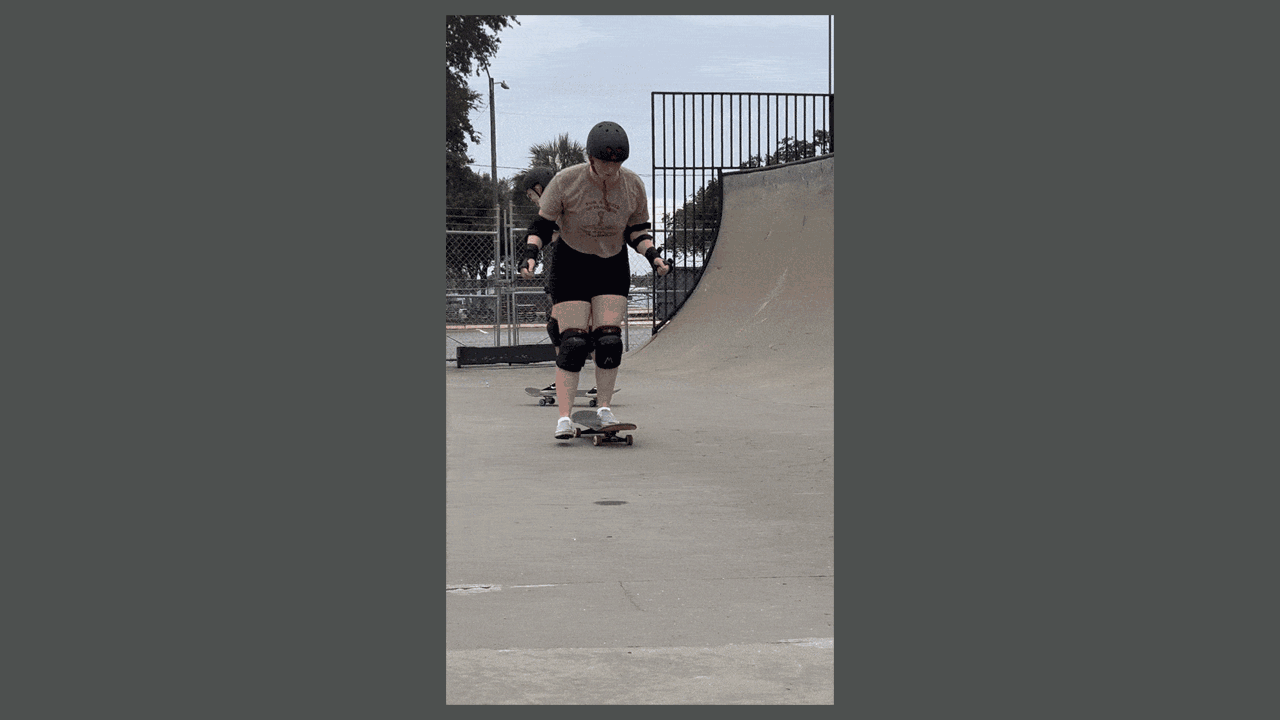 A GIF of a woman in black shorts in a brown T-shirt, wearing a helmet and knee, elbow and wrist pads tentatively pushes off the ground onto a skateboard.