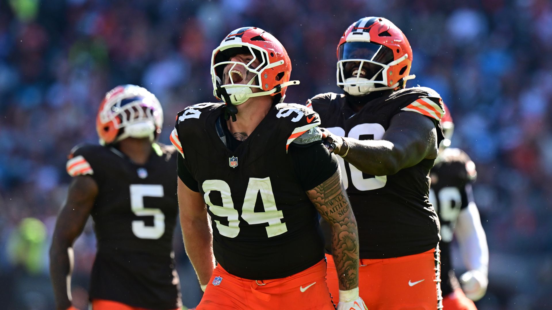 Two NFL players in orange pants and black jerseys celebrate on the field, embracing as the stadium crowd blurs in the background; orange helmets with white facemasks frame their faces.