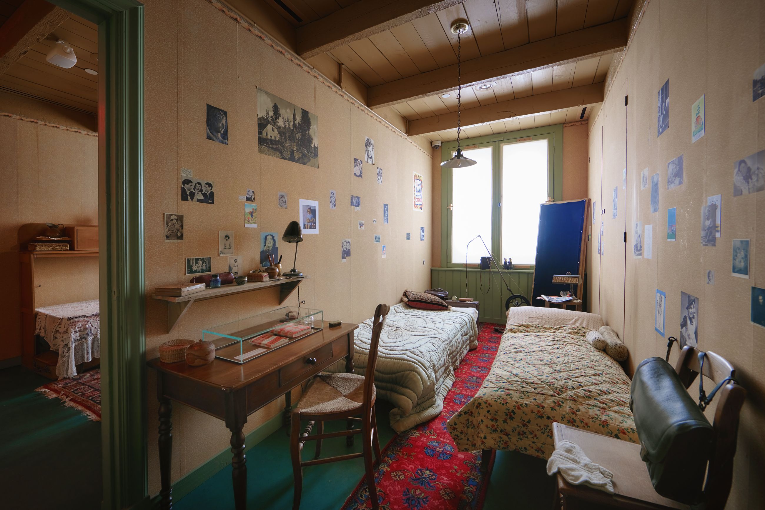 Long bedroom with beige walls covered in blue photos. Two beds along the right, a green door and window at the far end, a red floral rug, a hanging lamp, and a wooden desk with items.