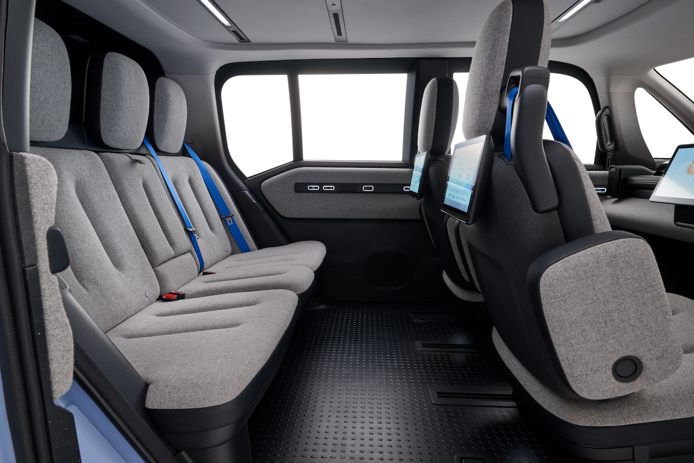 Interior of a modern vehicle with three rows of light gray fabric seats, blue seat belts, and tablet screens mounted on the back of the front seats; large windows give a bright, spacious cabin.