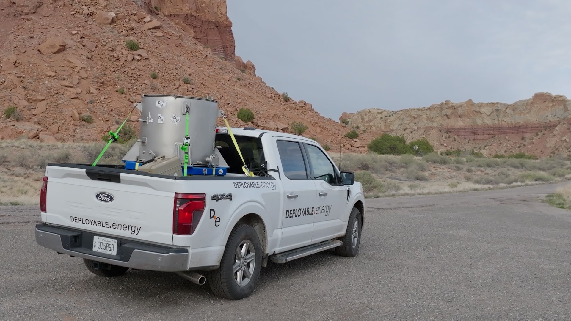 White Ford pickup with a large silver cylindrical tank in the bed, secured by bright green straps. Desert red rock cliffs in the background; the truck is branded "DEPLOYABLE.energy".
