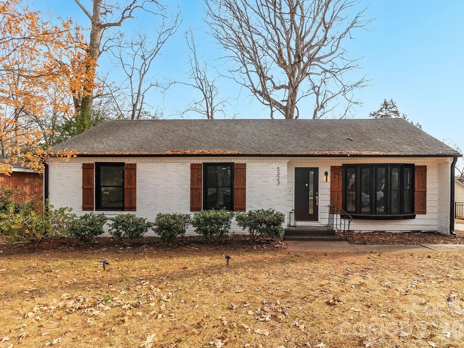 Single-story white brick house with brown wooden shutters, black windows, and a black front door, surrounded by shrubs and leaf-covered lawn under clear blue sky in autumn.