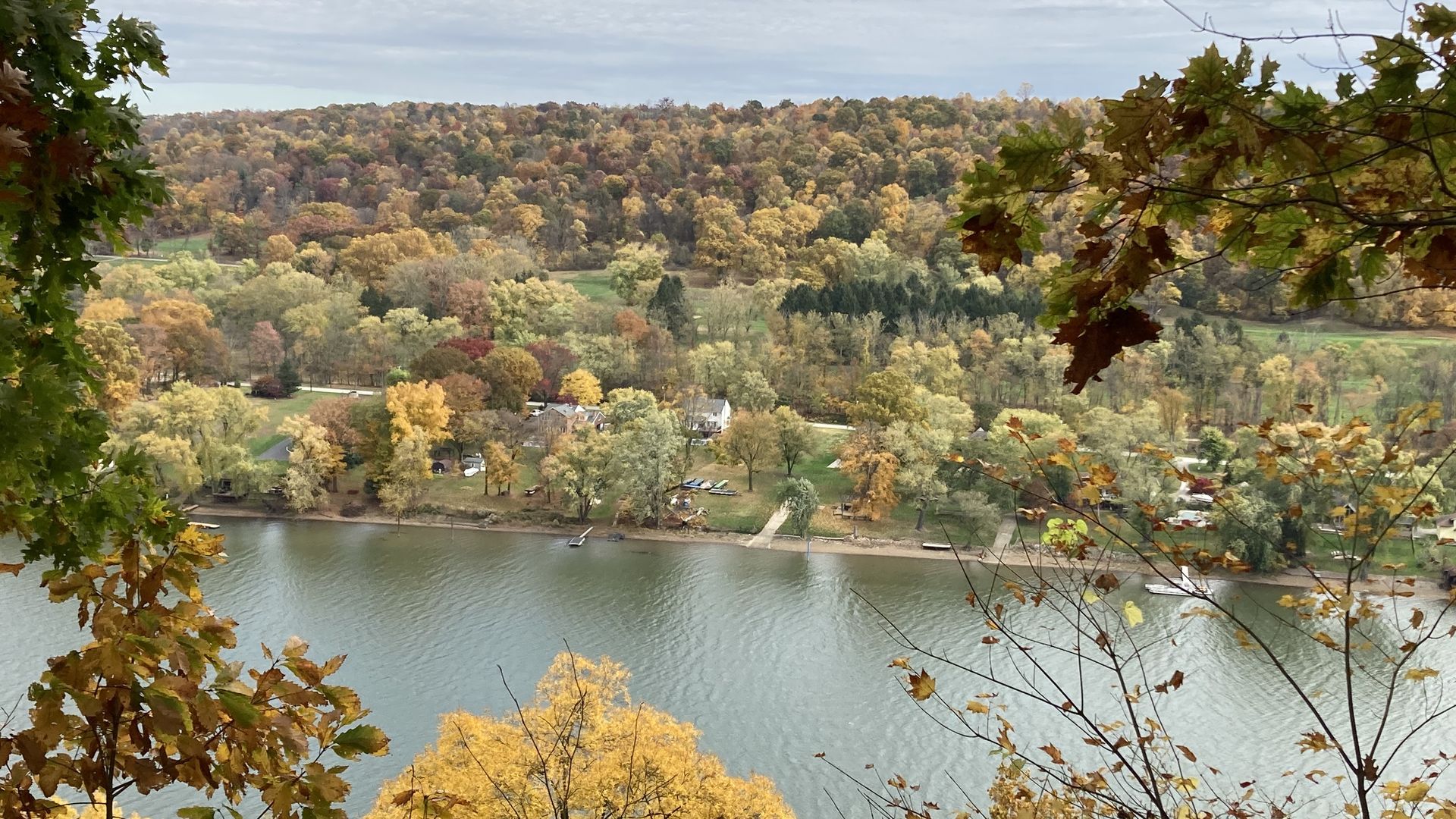 A river flows through a landscape of trees in fall colors, with shades of green, yellow, orange, and red. Houses and docks line the riverbank under a cloudy sky.