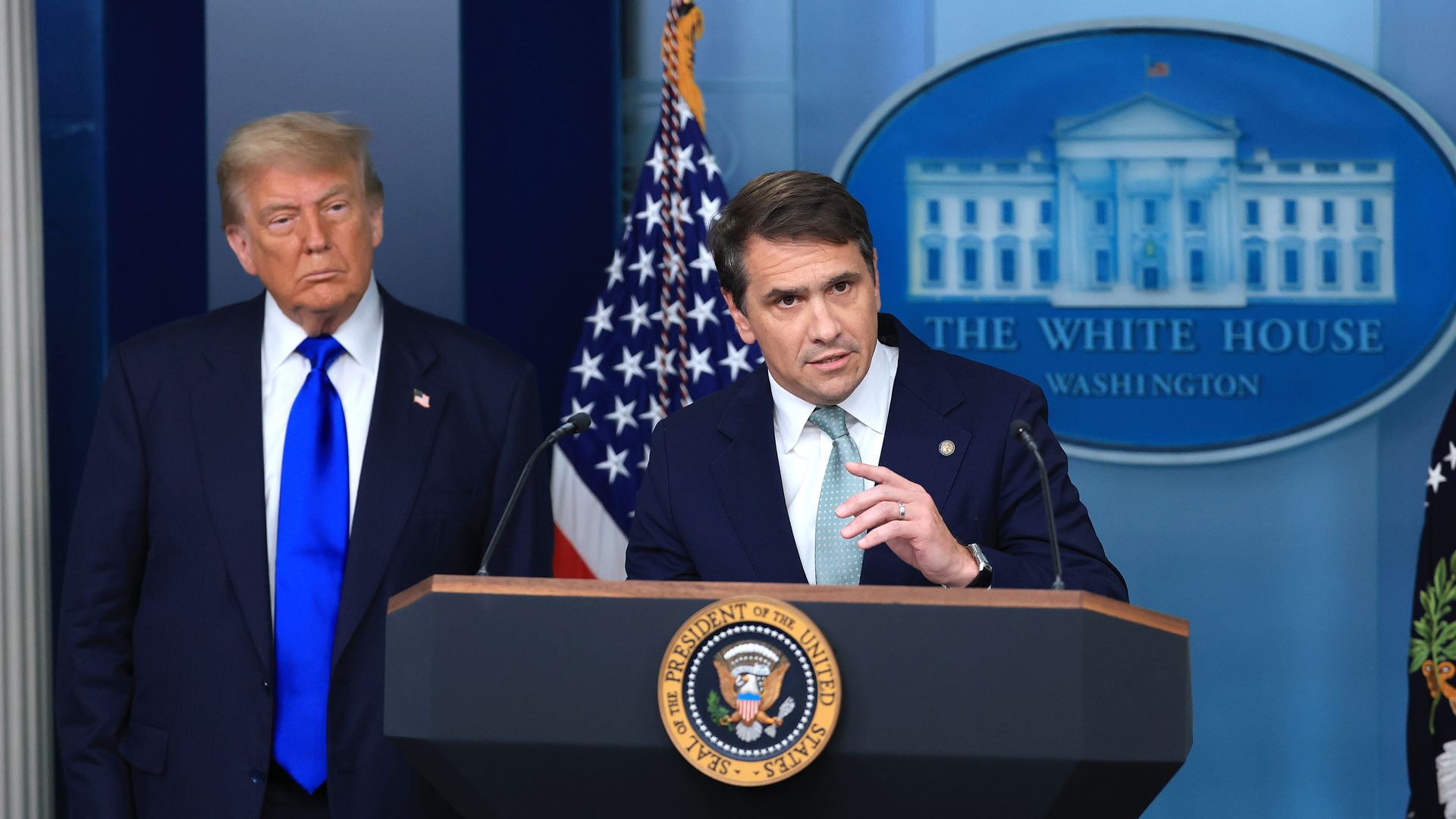 President Trump and U.S. Deputy Attorney General Todd Blanche in dark suits stand at a podium with the presidential seal; one speaks into a microphone while the other stands to the left. American flag and a White House backdrop behind them.