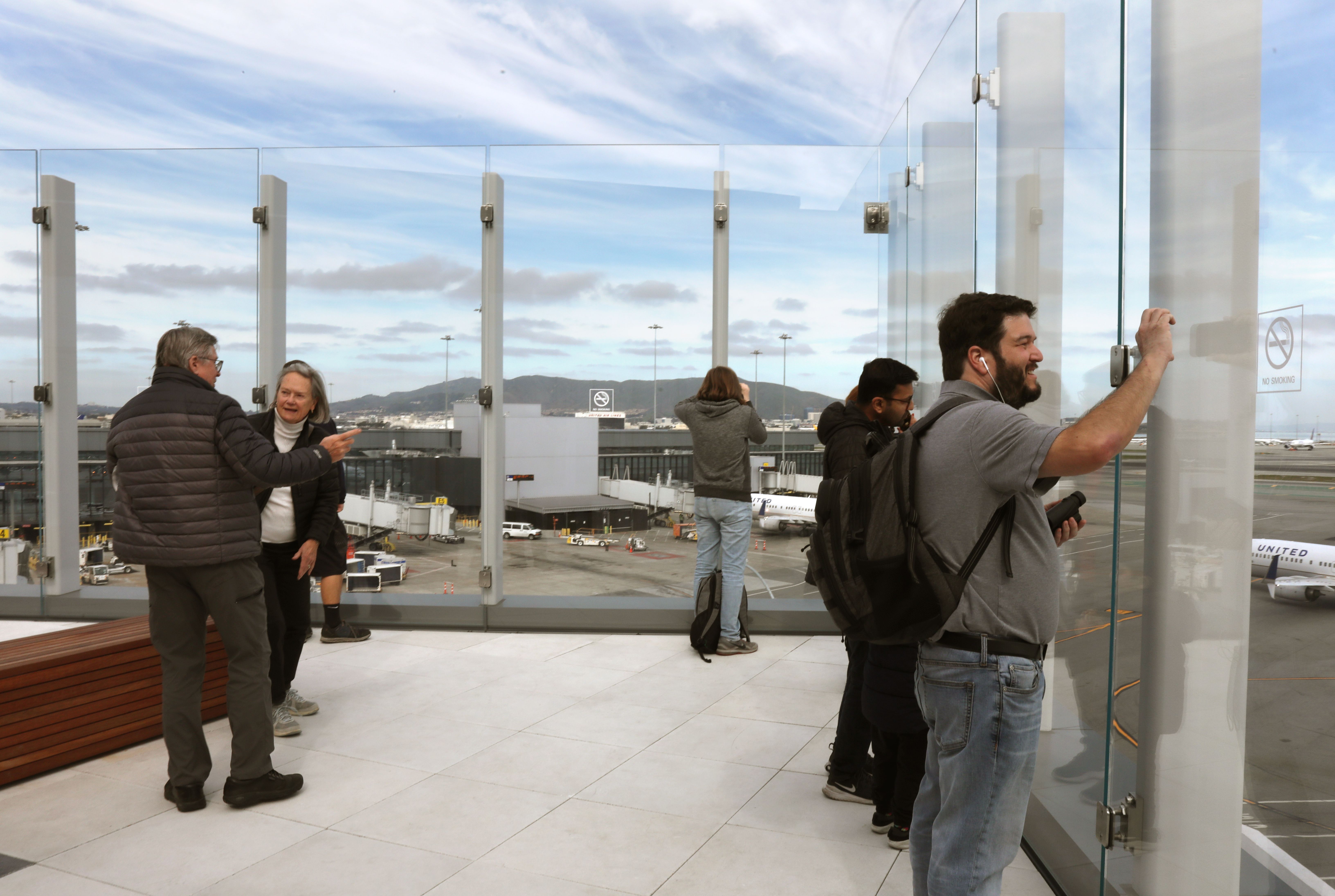 People at an airport observation deck with glass barriers looking at planes and the runway under a partly cloudy sky.