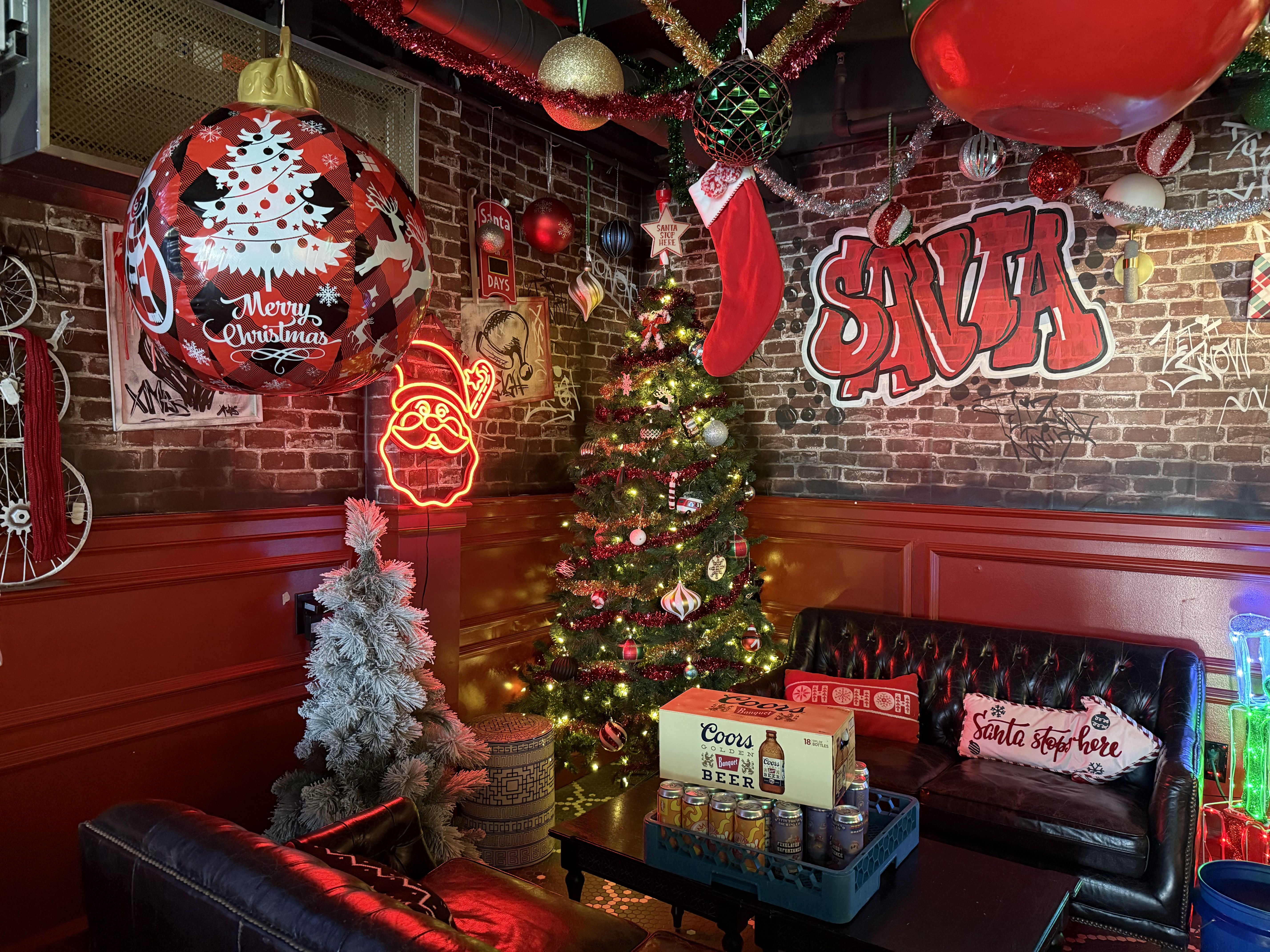 Cozy room with a decorated Christmas tree, red and black ornaments, a red stocking, neon Santa face, and graffiti-style "SANTA" on brick wall. Black leather couches with festive pillows.