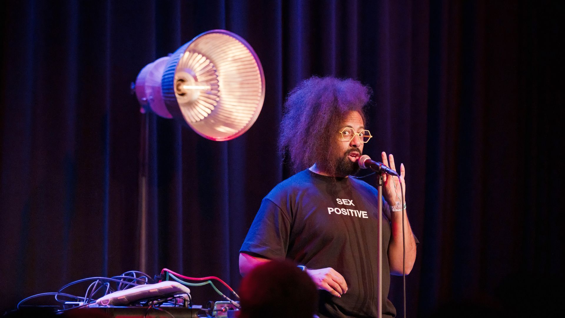 A dimily lit photo of comedian and musician Reggie Walks talking into a mic on stage as he does a set in Berlin, wearing glasses and a black T-shirt that says "sex positive."