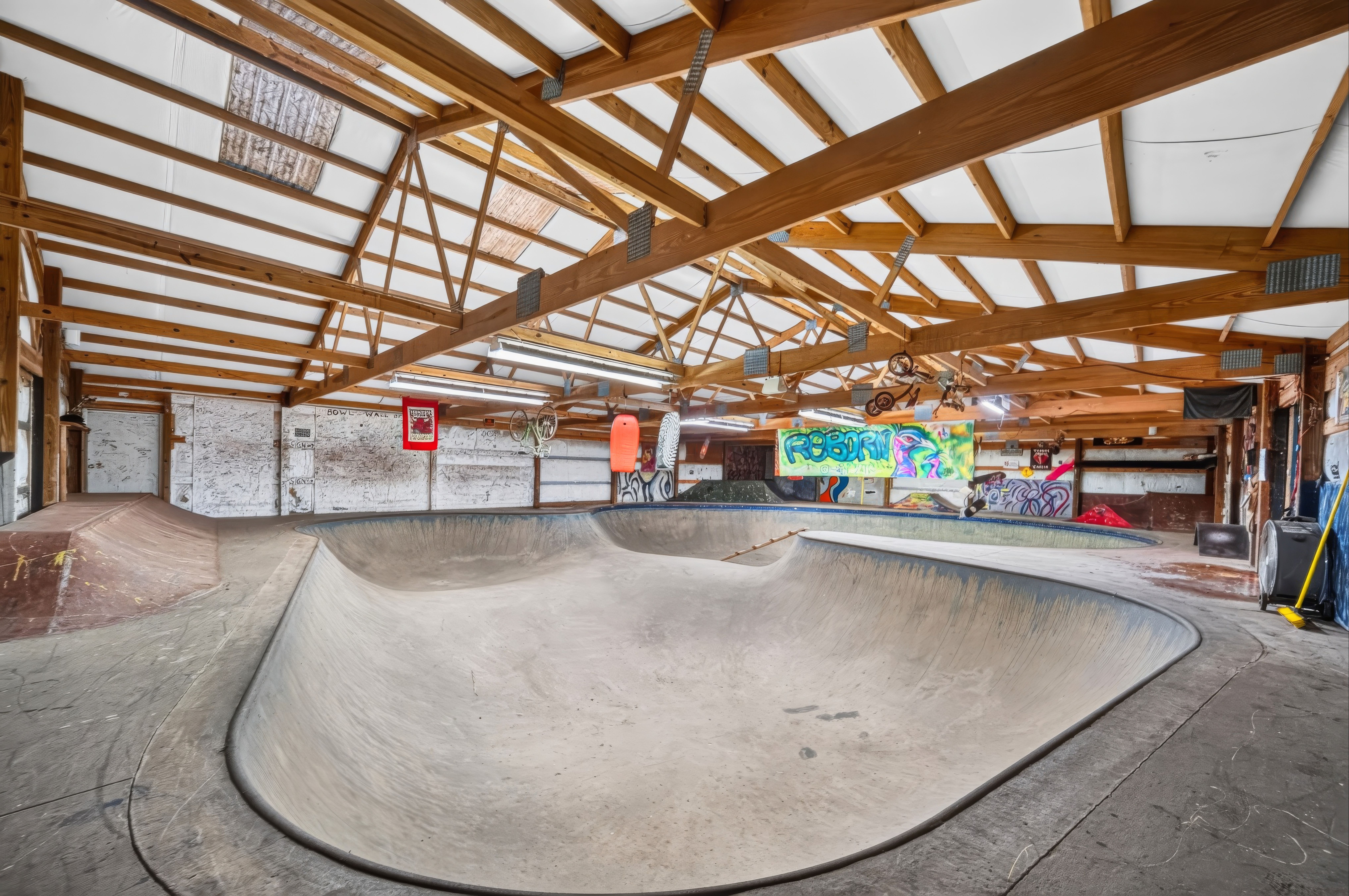 Indoor skatepark with a timber beam ceiling and white panels. A large curved concrete bowl dominates the foreground, with graffiti walls and hanging banners in the background.