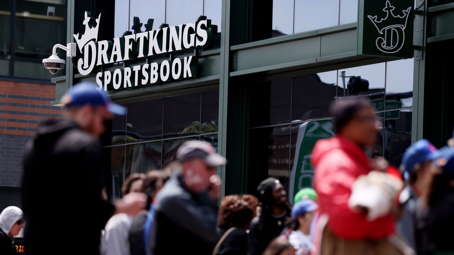 DraftKings Sportsbook storefront with a white crown logo and bold sign on glass; reflective windows. A blurred crowd walks by as a security camera sits left.