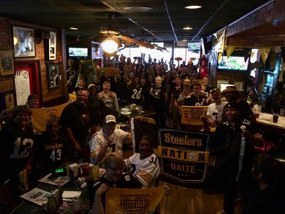 A large group of Steelers fans are assembled in a bar setting. They're holding Terrible Towels and have a "Steelers Nation Unite" banner at the front of the group.