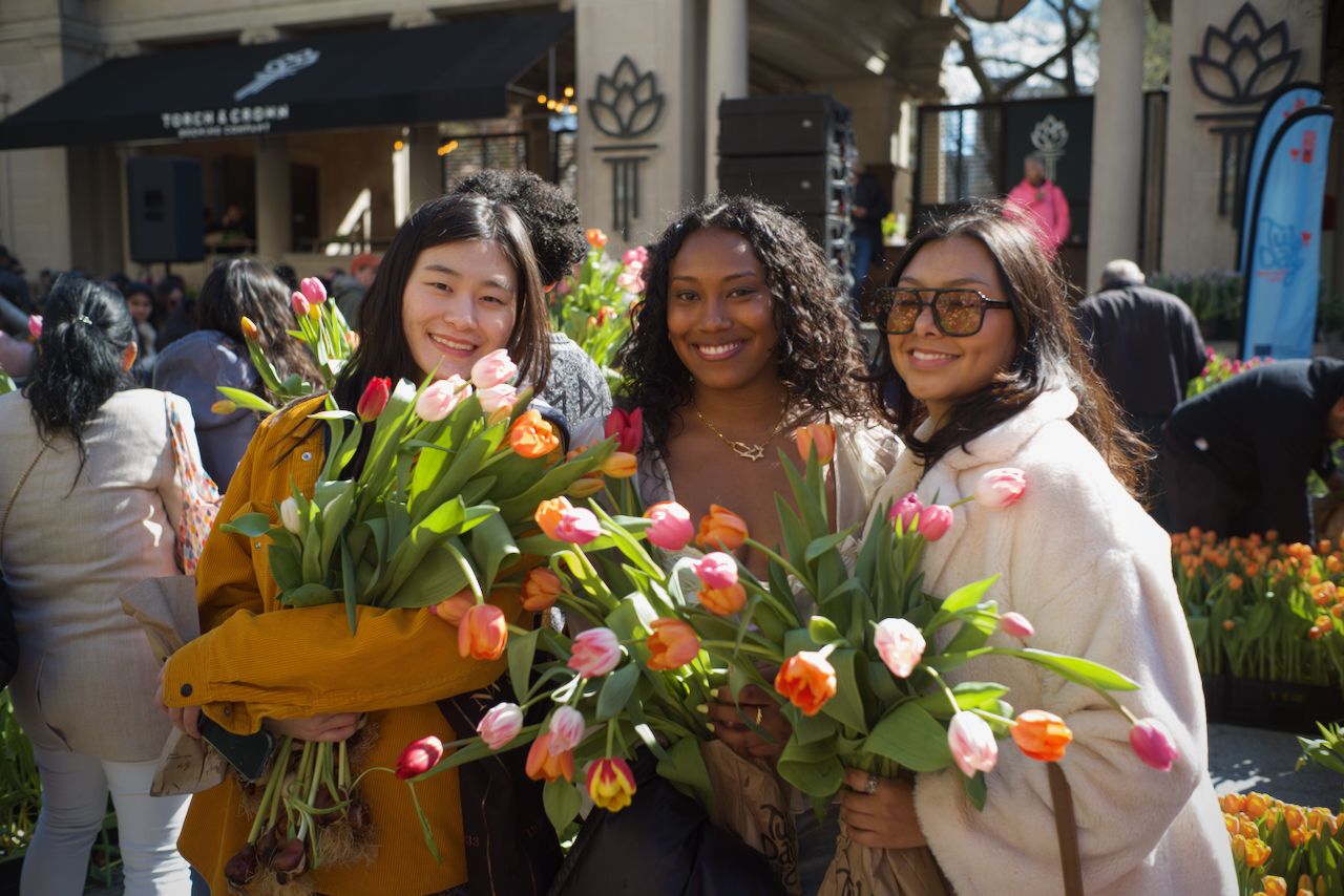 Three women smiling outdoors holding colorful tulip bouquets, one in a yellow jacket, one with curly hair, and one wearing large sunglasses and a white coat, surrounded by more flowers.
