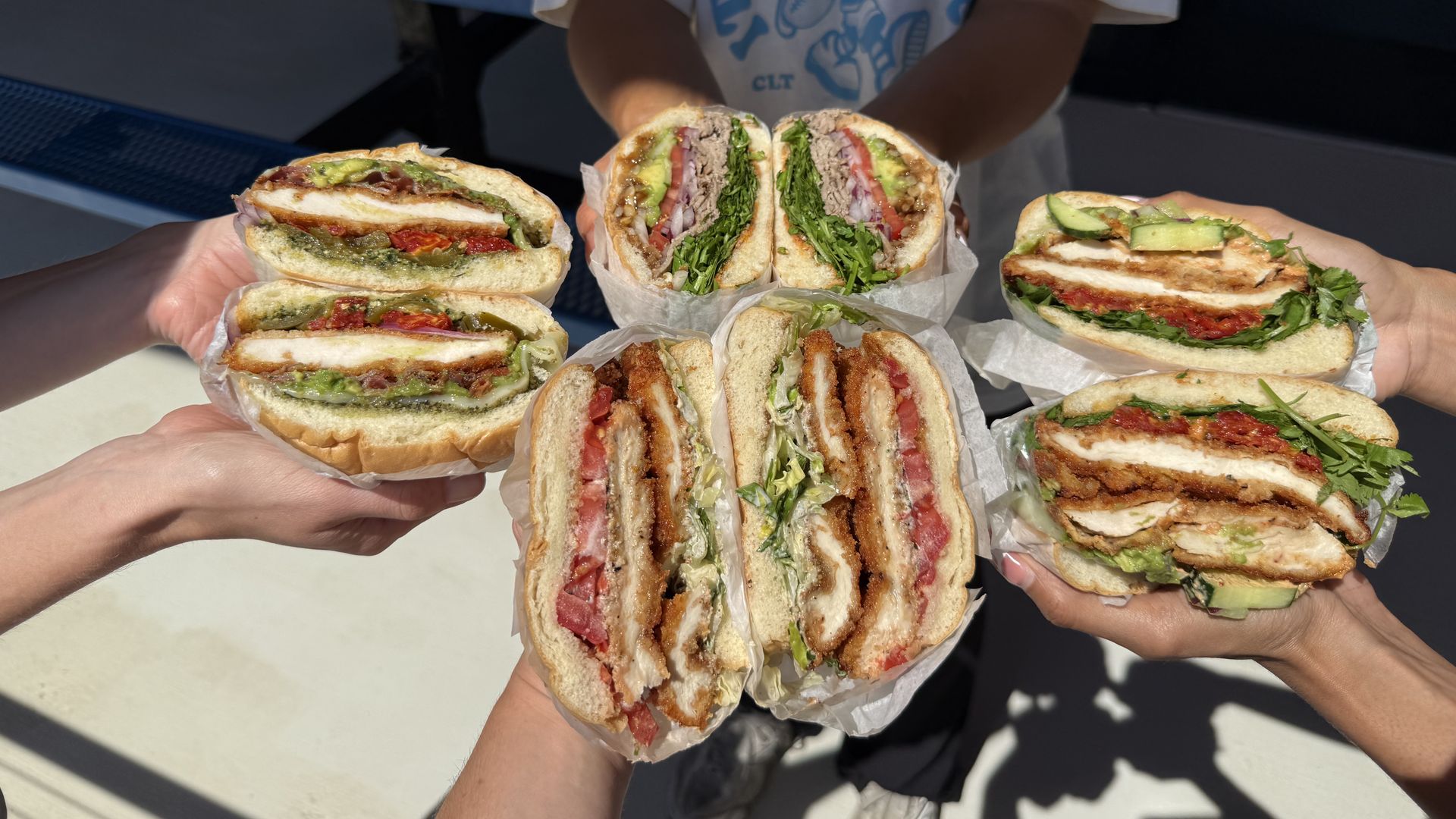 People extend many sandwich halves toward the camera; fillings include lettuce, tomato, avocado, onions, and meat. A person in a light blue graphic shirt stands in the background outdoors.