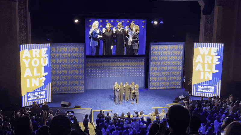 A stage event with several people standing on stage, seen on a large screen above them. Audience watches. Signs on sides display "ARE YOU ALL IN?" and "WE ARE ALL IN" with "ALL IN FOR NEW ORLEANS".