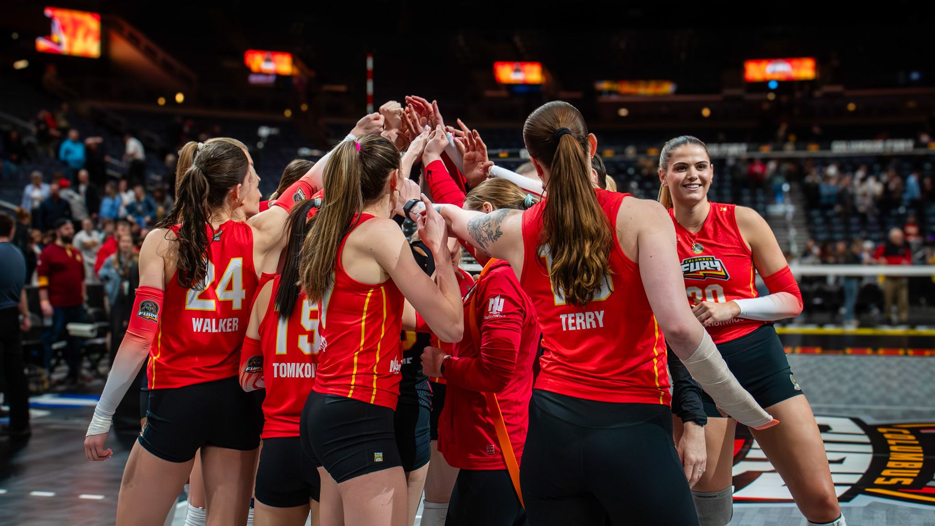 Columbus Fury women's volleyball team in red jerseys huddle with hands together on court, with players named Walker, Tomko, and Terry visible, and a player smiling in the background.