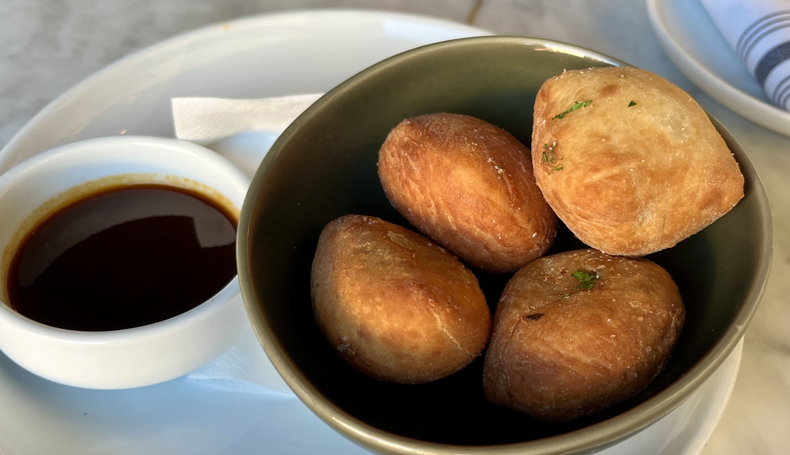 Four golden brown fried pastries in a green bowl with a small white cup of dark dipping sauce on a white plate, accompanied by a white napkin on a marble surface.