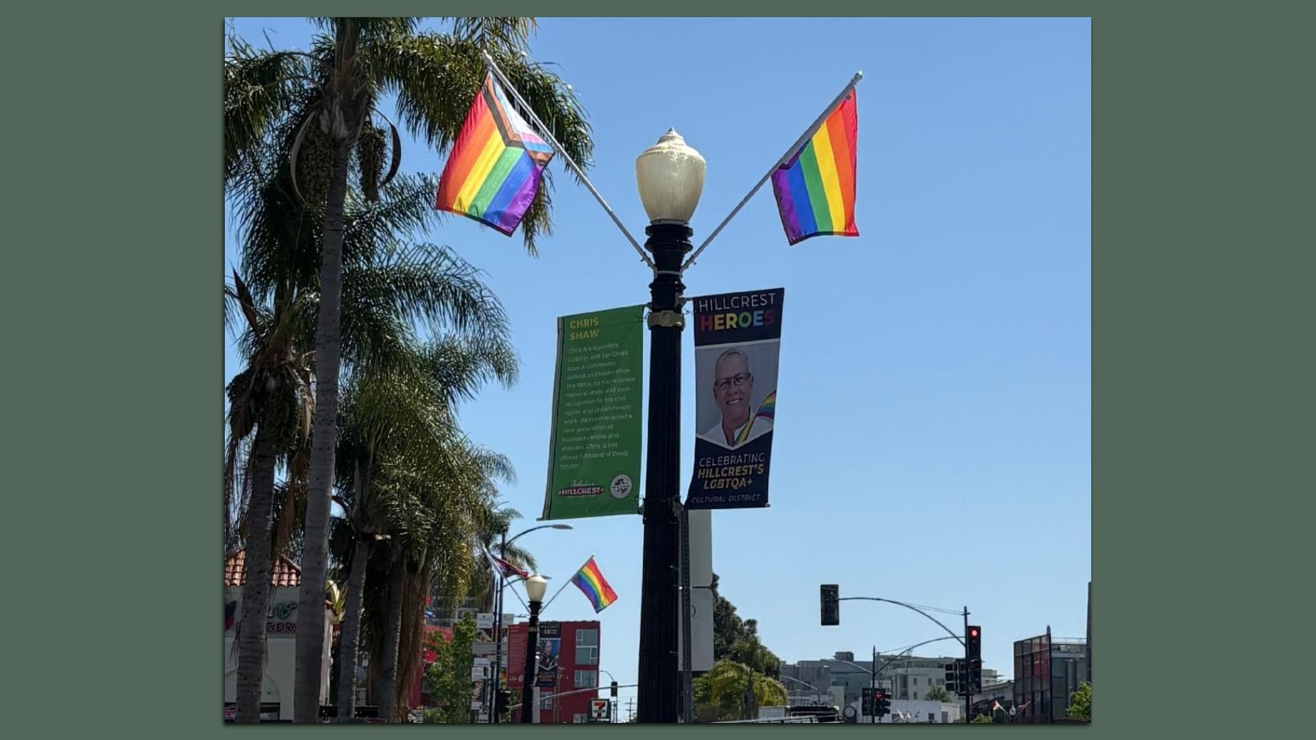Sunlit urban street scene with palm trees and a black lamppost adorned by two rainbow Pride flags; banners for Hillcrest Heroes hang from the post against a clear blue sky.