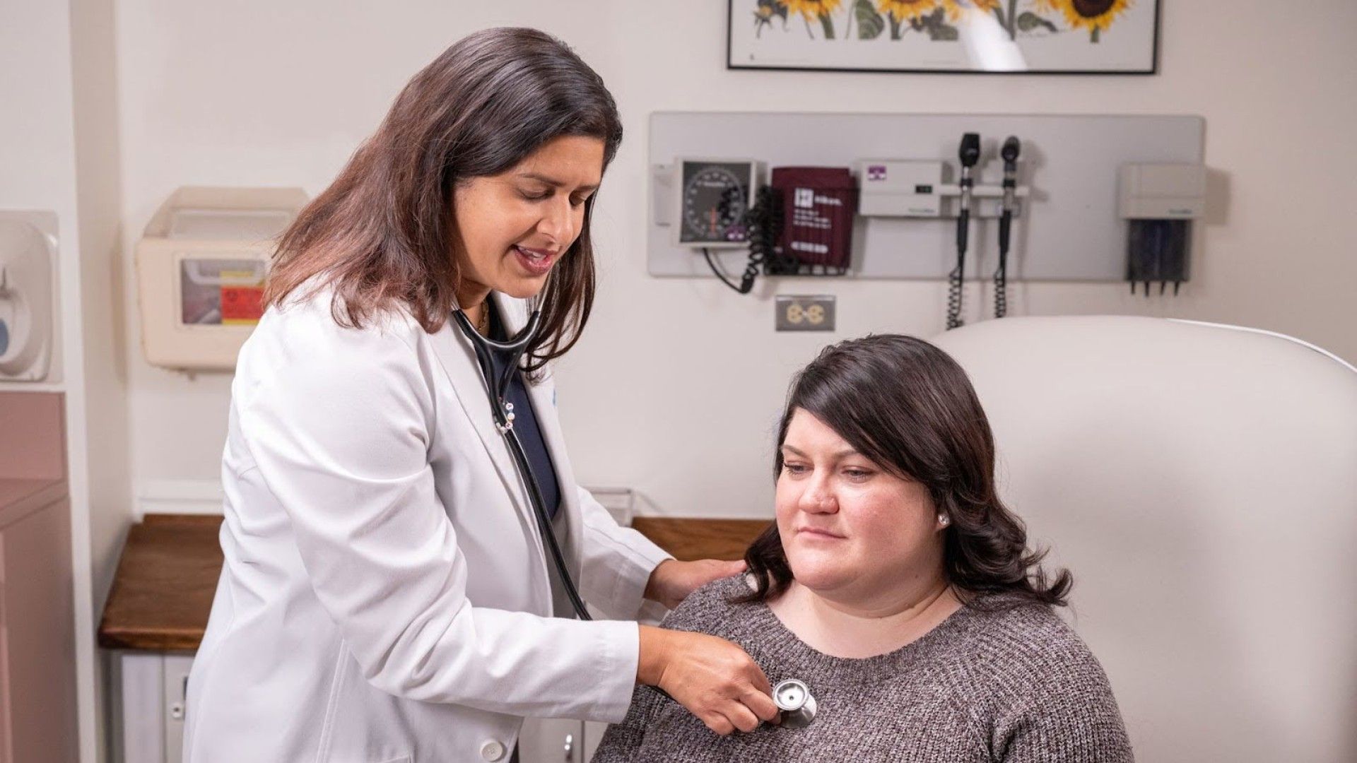 A female doctor in a white coat uses a stethoscope to examine a seated patient wearing a gray sweater in a medical exam room with equipment on the wall and a sunflower poster.