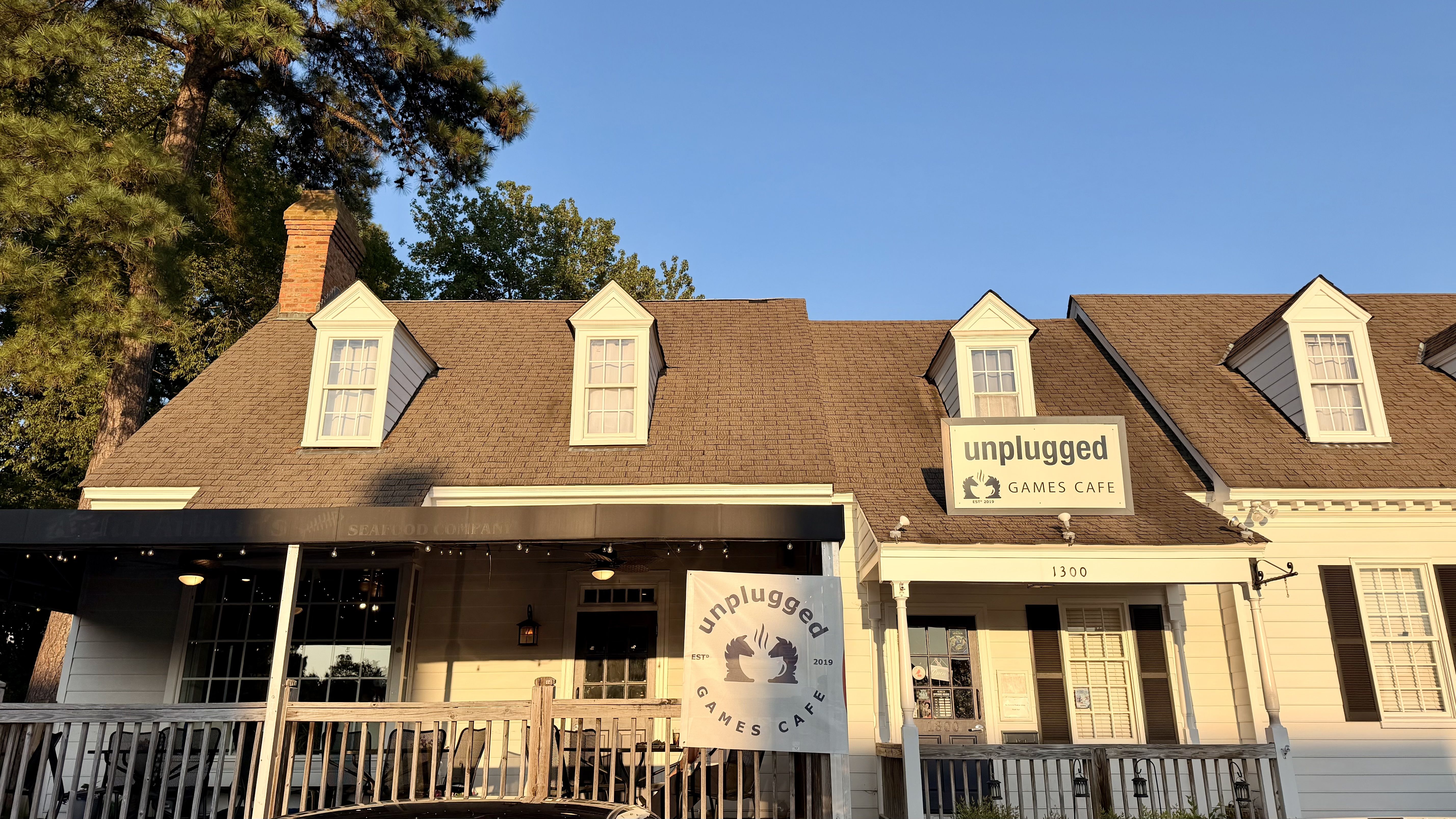 Sunlit storefront with a brown shingle roof, three white dormer windows, and a brick chimney. A sign and banner read "unplugged GAMES CAFE" beneath a clear blue sky with trees on the left.