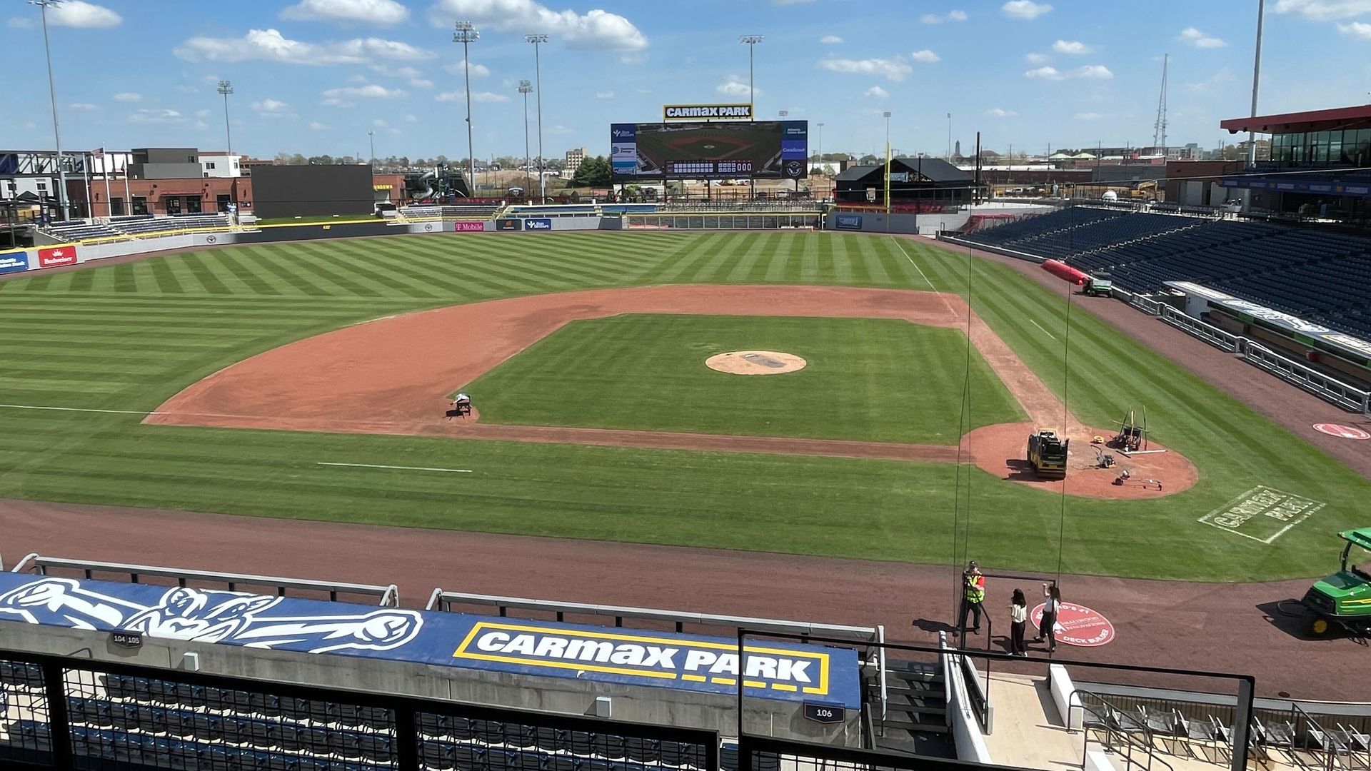 Wide view of a baseball stadium under a bright blue sky with scattered clouds. Green field with a dirt infield, empty blue seats, tall light towers, and a scoreboard reading "CARMAX PARK".