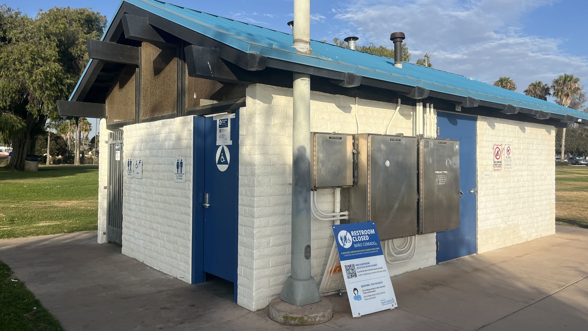 White brick building with a bright blue metal roof and black trim. Exposed pipes and panels, a gray street lamp out front, and a "Restroom Closed" sign. Blue doors; trees and blue sky behind.