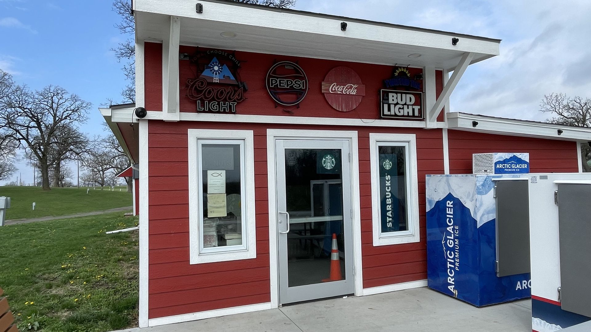 Red kiosk with white trim and signs for Coors Light, Pepsi, Coca-Cola, and Bud Light above the door. A blue Arctic Glacier ice machine outside; green lawn and bare trees in the background.