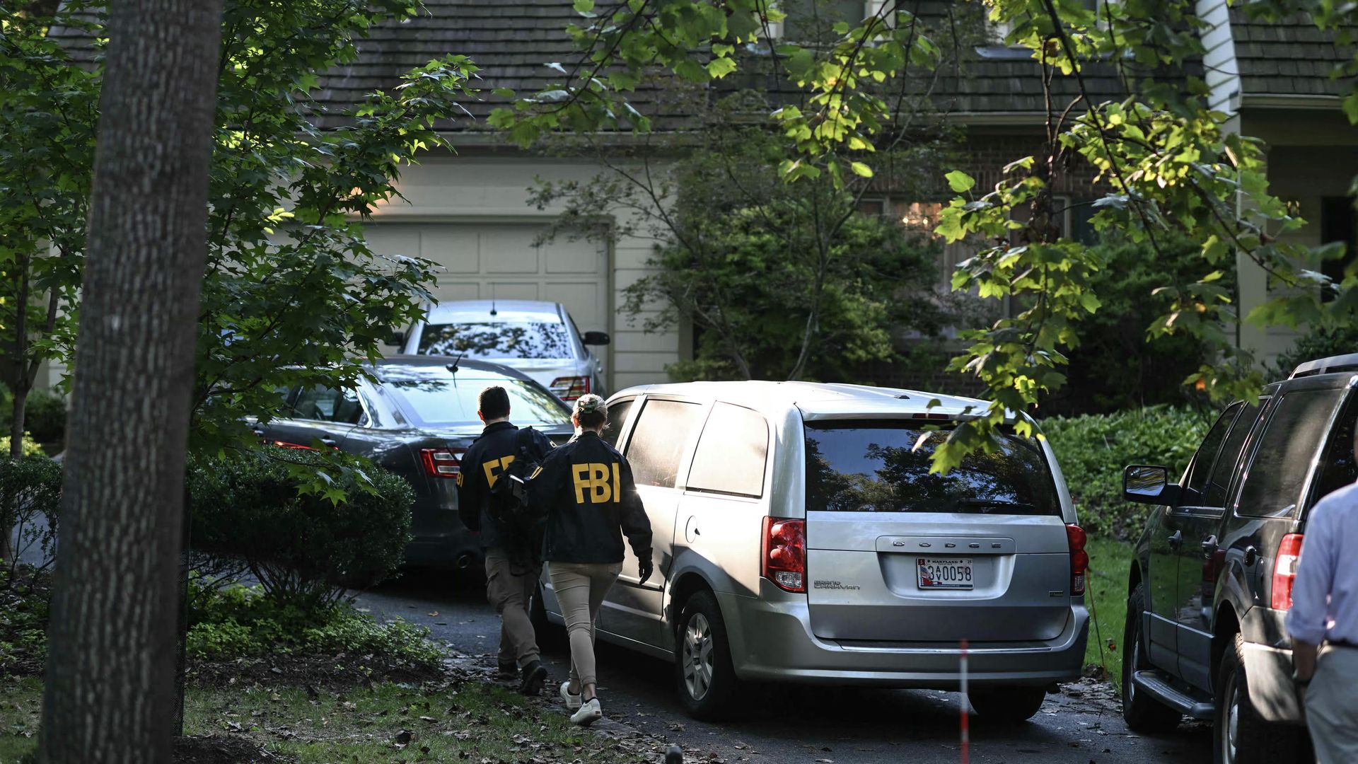 Two FBI agents wearing black jackets with yellow letters walk towards a suburban house driveway with several parked cars and green trees around.