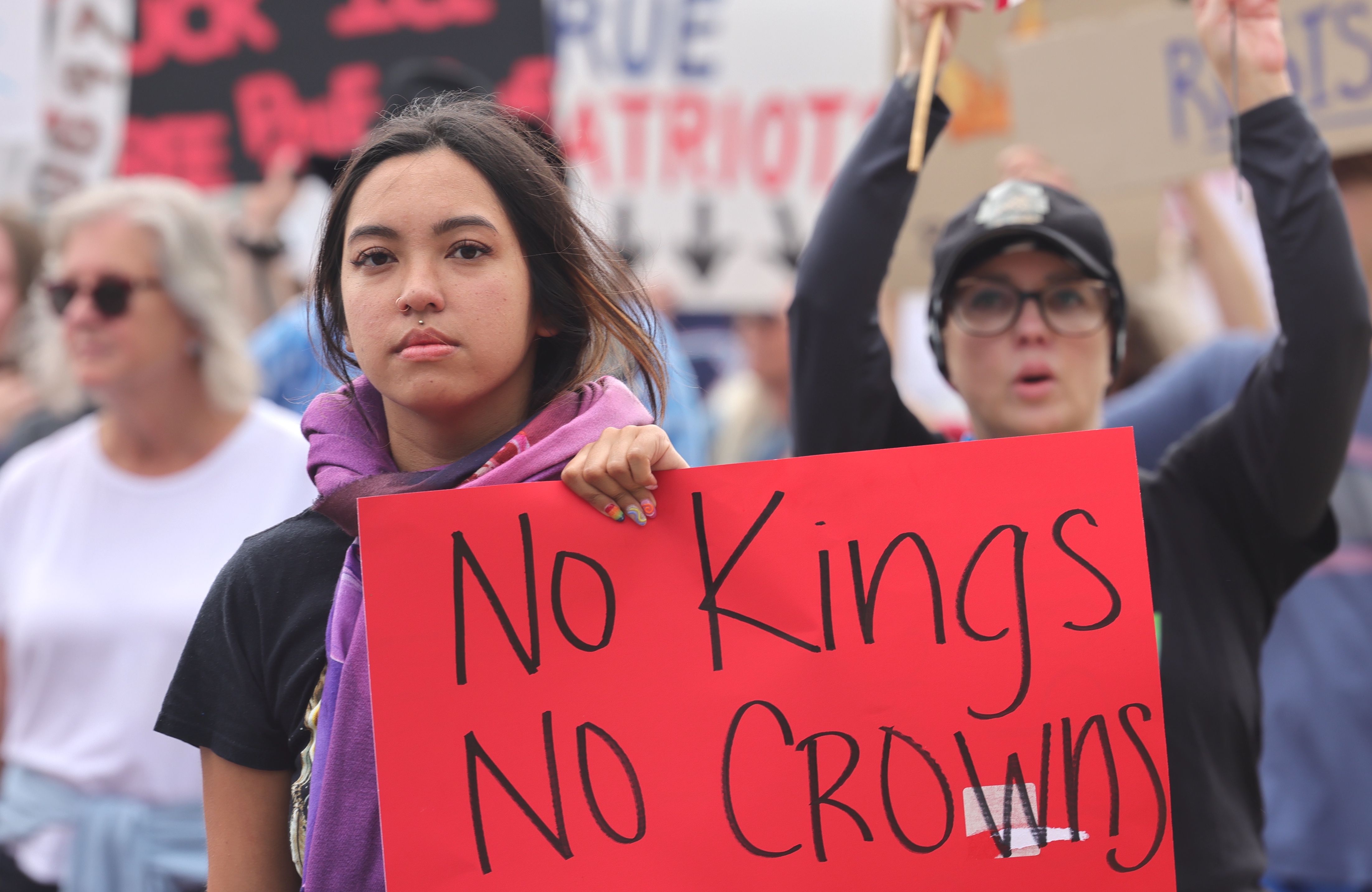 Woman with a purple scarf holds a red sign reading "No Kings No Crowns" at a protest, surrounded by other demonstrators.