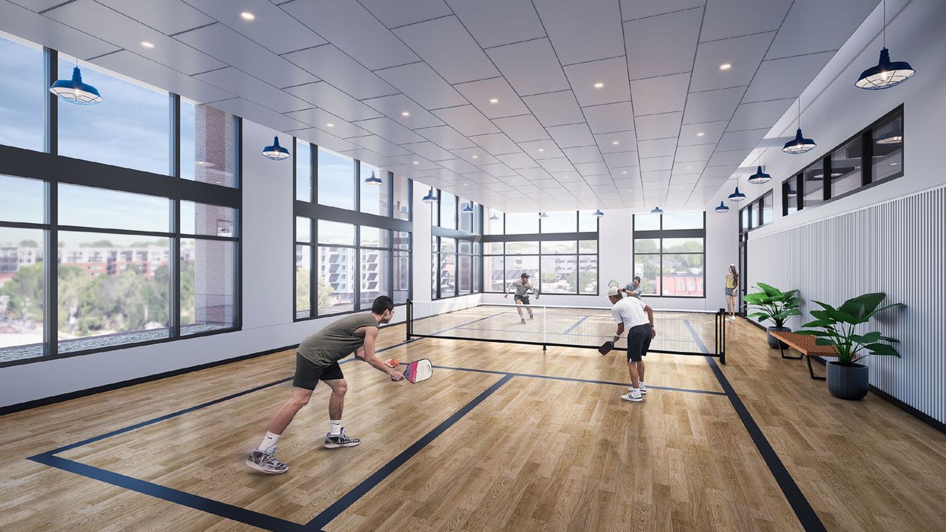 Four people playing pickleball indoors on a wooden court with large windows, blue lights, and potted plants in a bright, modern room with white walls and ceiling.