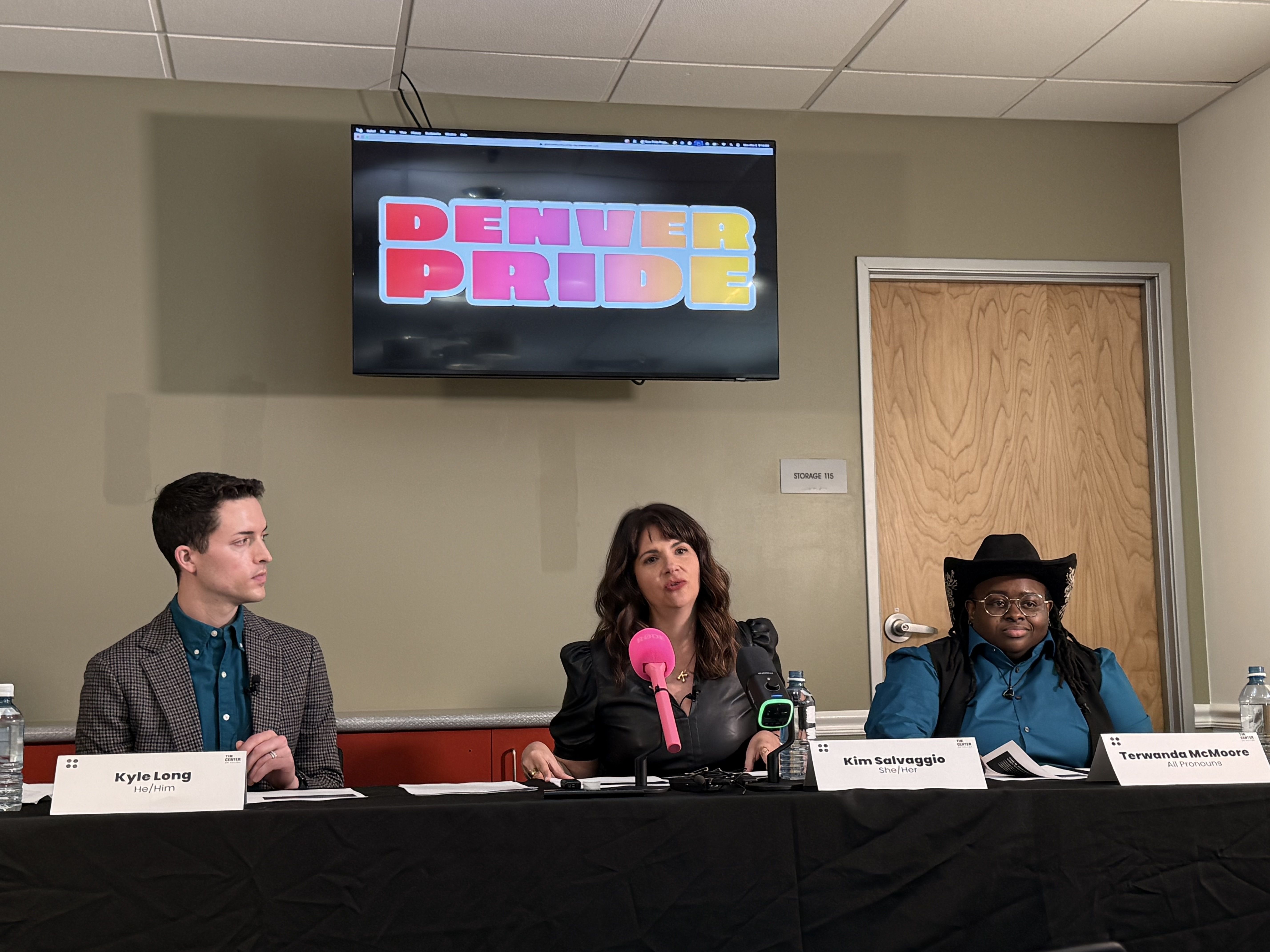 Three panelists at a Denver Pride event, seated behind a black table with microphones. Screen above displays colorful "DENVER PRIDE" text in red, purple, yellow, and orange.