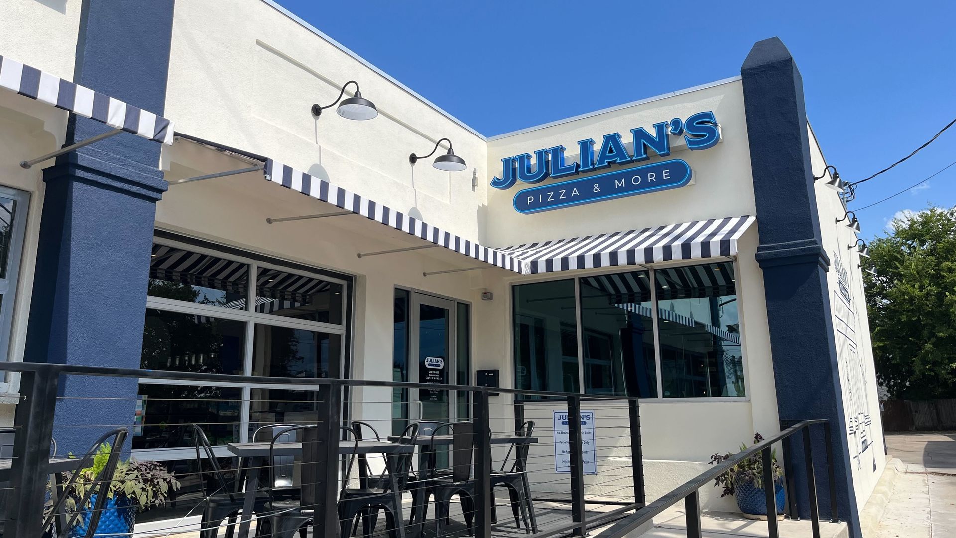 Exterior of Julian's Pizza & More with blue and white striped awnings, black metal chairs and tables on patio, white and blue building, and clear blue sky above.