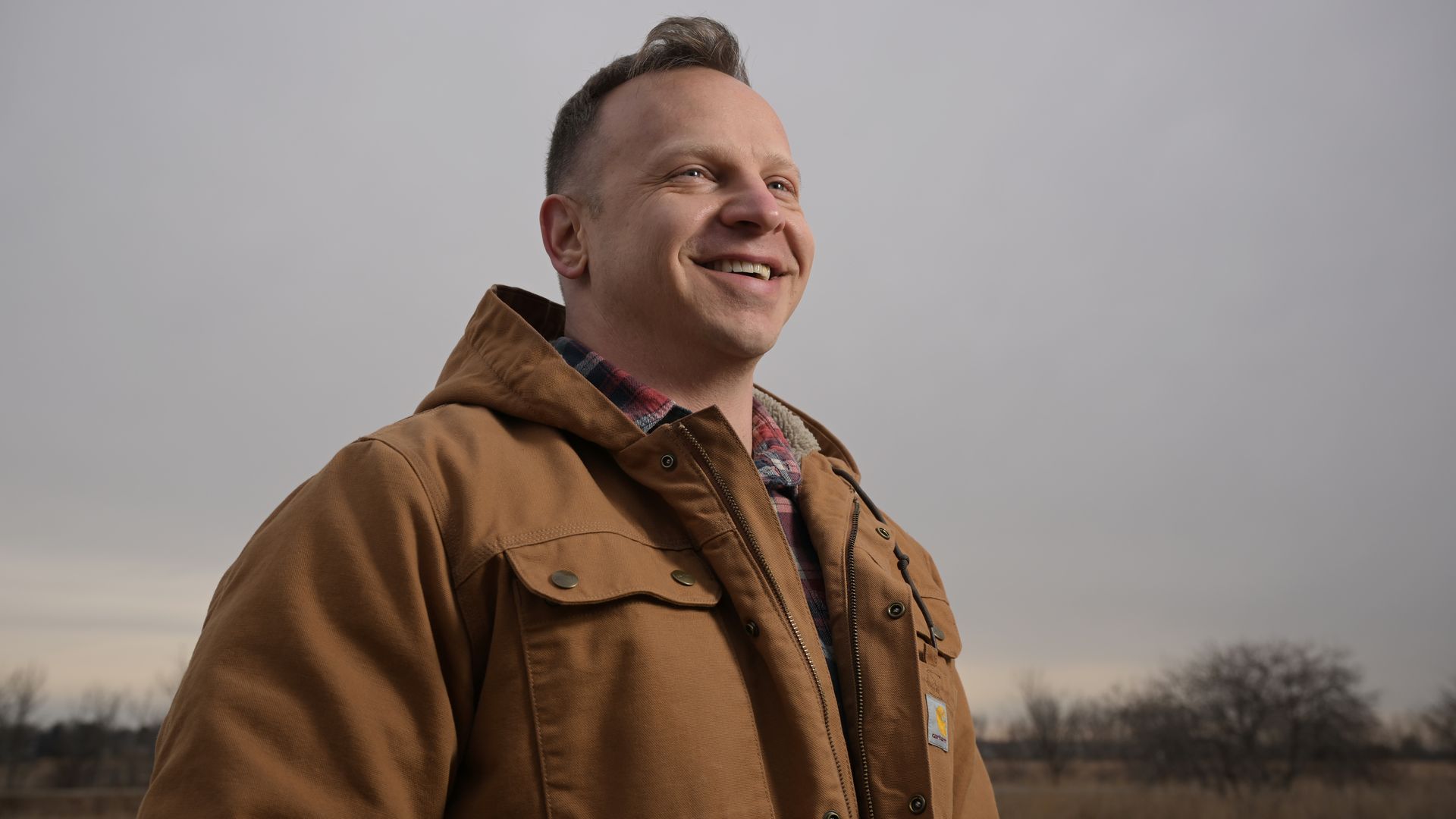 Smiling man wearing a tan jacket over a plaid shirt stands outdoors against a gray cloudy sky, with a distant field and bare trees in view.