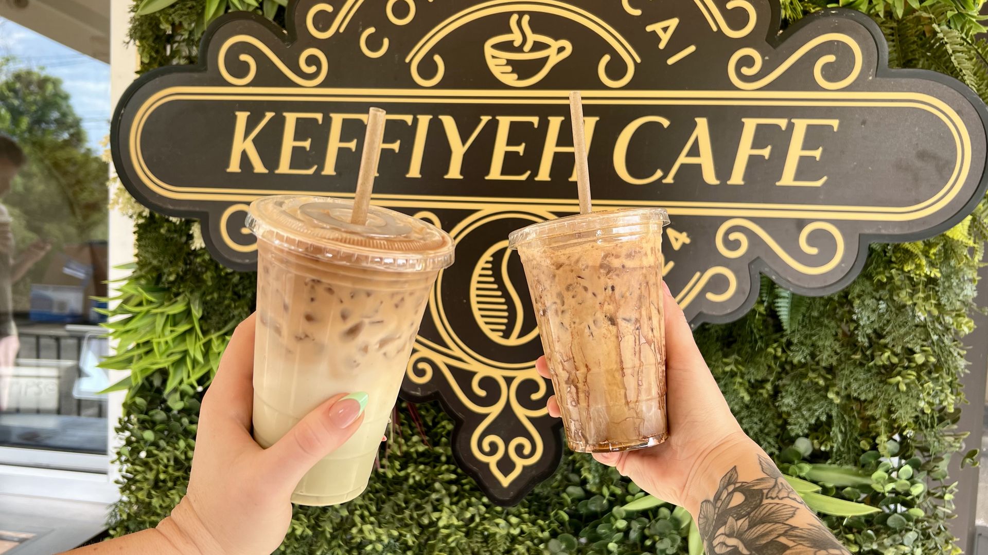 two hands each holding up a latte in front of a sign that says "keffiyeh cafe" which is written in gold letters