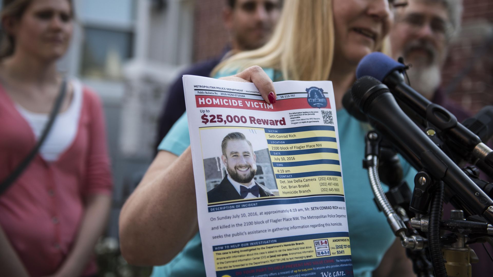 Photo of Mary Rich speaking into a mic and holding a flyer asking for information on Seth Rich's death