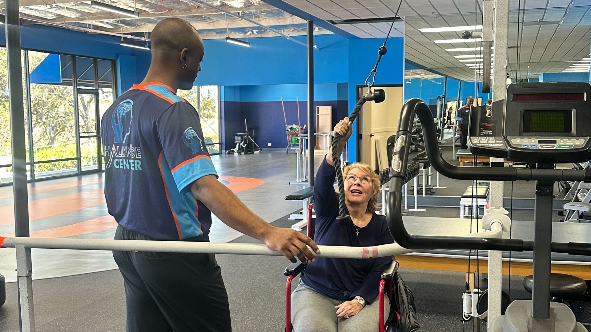 Trainer in a blue shirt assists a woman in a red wheelchair as she pulls a pulley cord for an upper-body exercise in a bright gym, with blue walls and treadmills in the background.