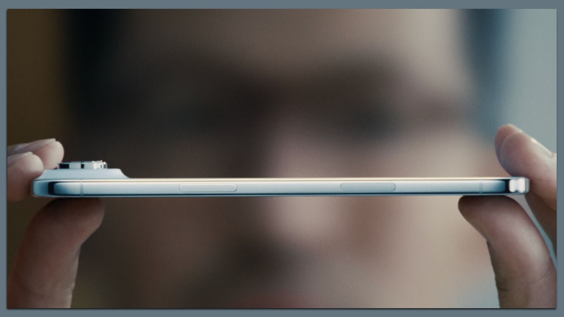 Close-up of a person holding a slim, silver smartphone horizontally, showing its thin side with volume and power buttons against a blurred background.