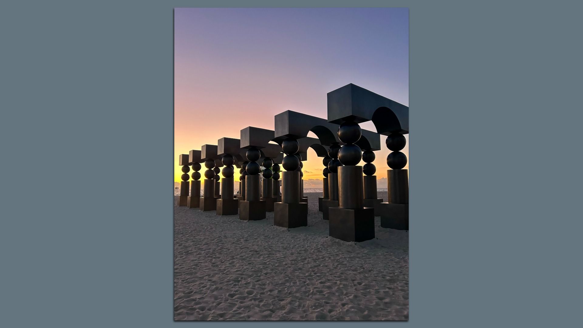 Black geometric sculptures in rows on sandy beach during a colorful sunrise with purple and orange sky.