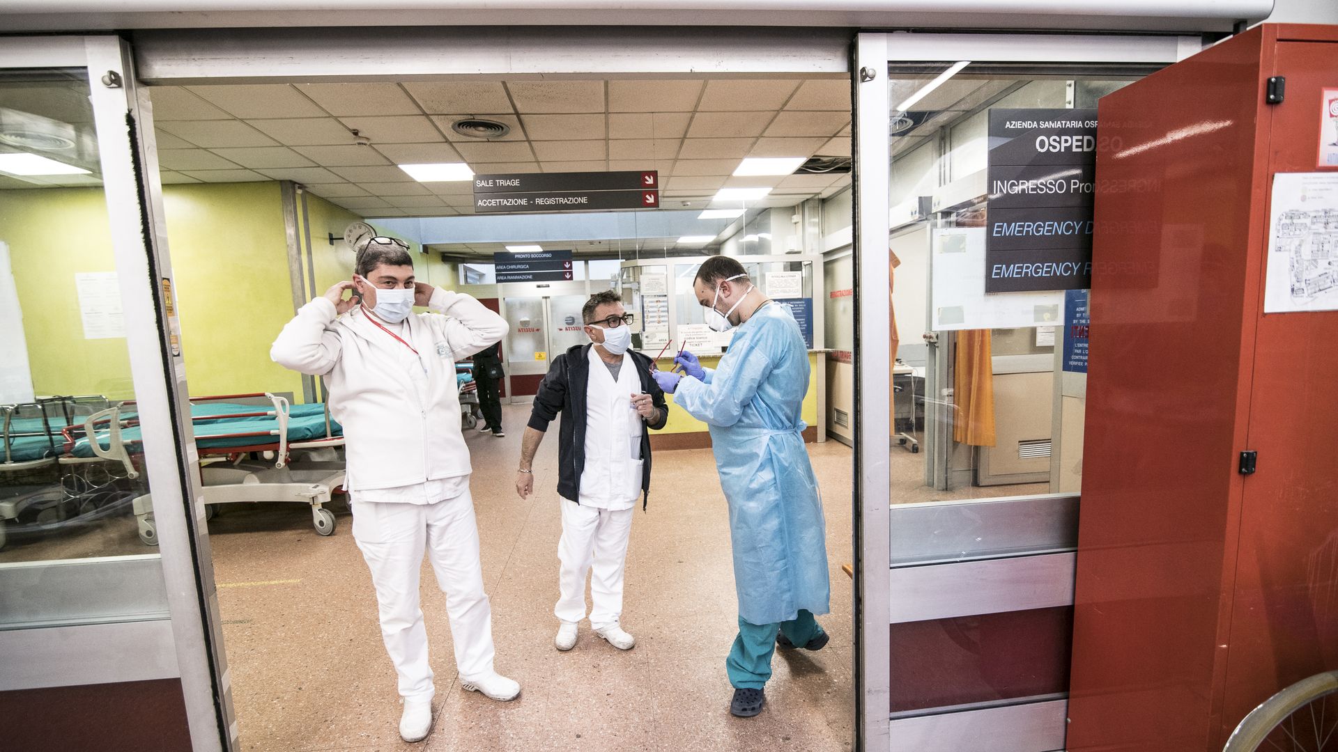 Three fully masked medical workers stand inside the doors of a hospital ER.