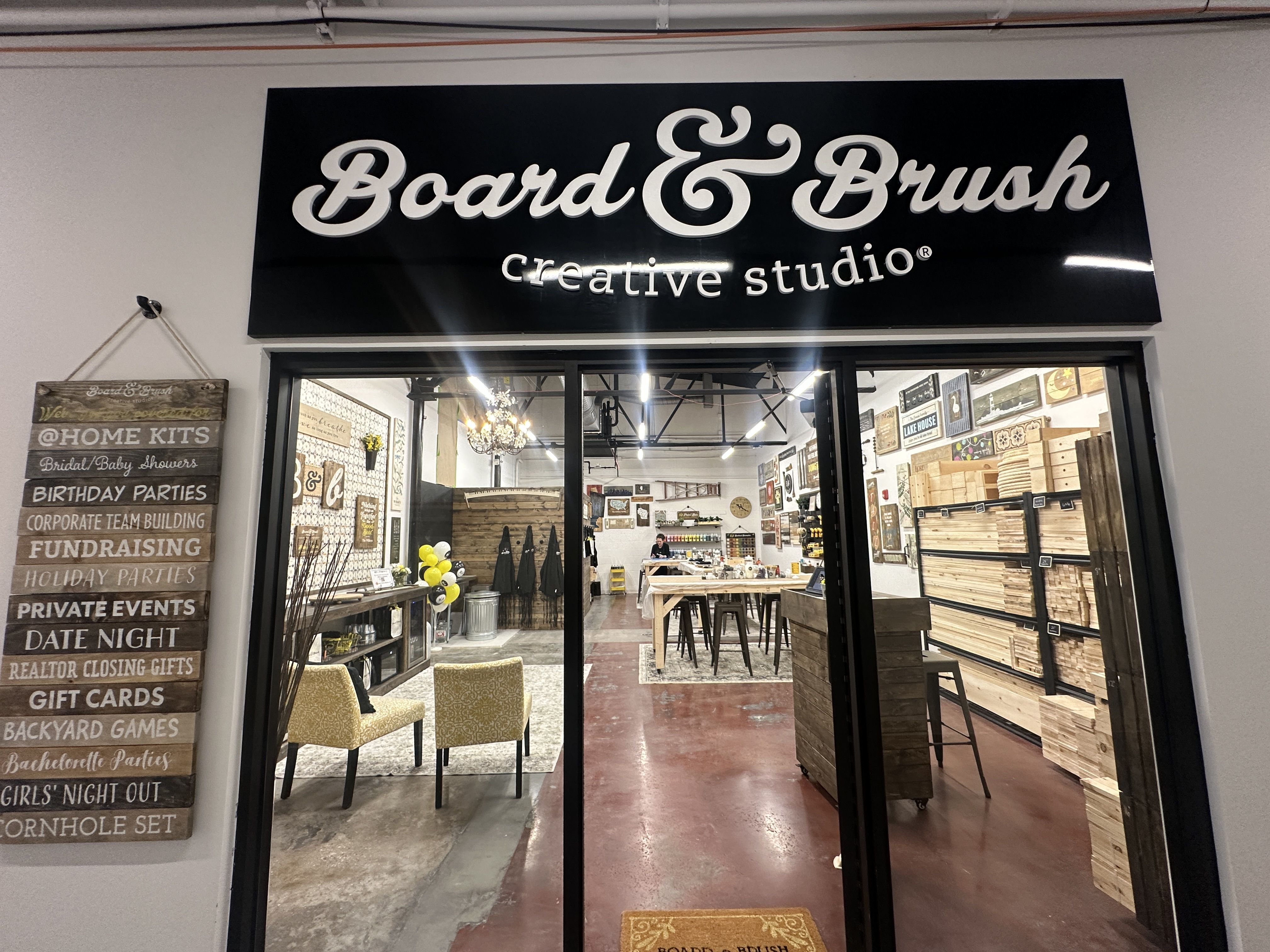 Board & Brush. Front of a Board & Brush creative studio with open glass doors. Inside is a rustic craft workshop: wooden planks on shelves, stools at a central worktable, and decorative signs on walls.