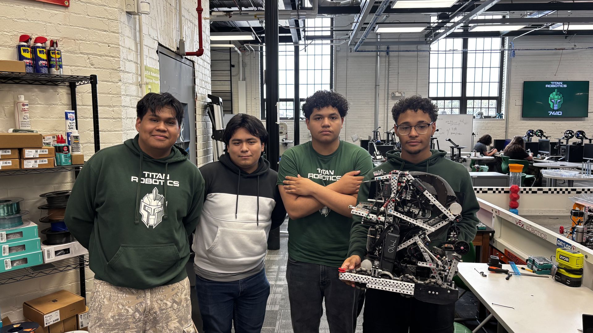 Four students in a robotics lab pose for a photo; three wear green "Titan Robotics" hoodies, the fourth holds a large robot frame. Background shows shelves and a Titan Robotics screen.