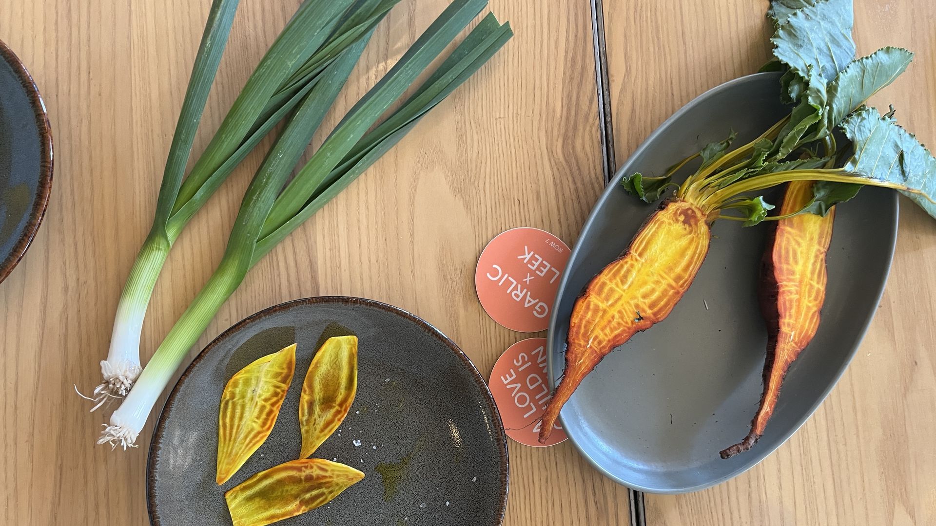 Two yellow-orange carrots with green tops on a gray plate, three carrot slices on a smaller gray plate, and two green onions on a wooden table.