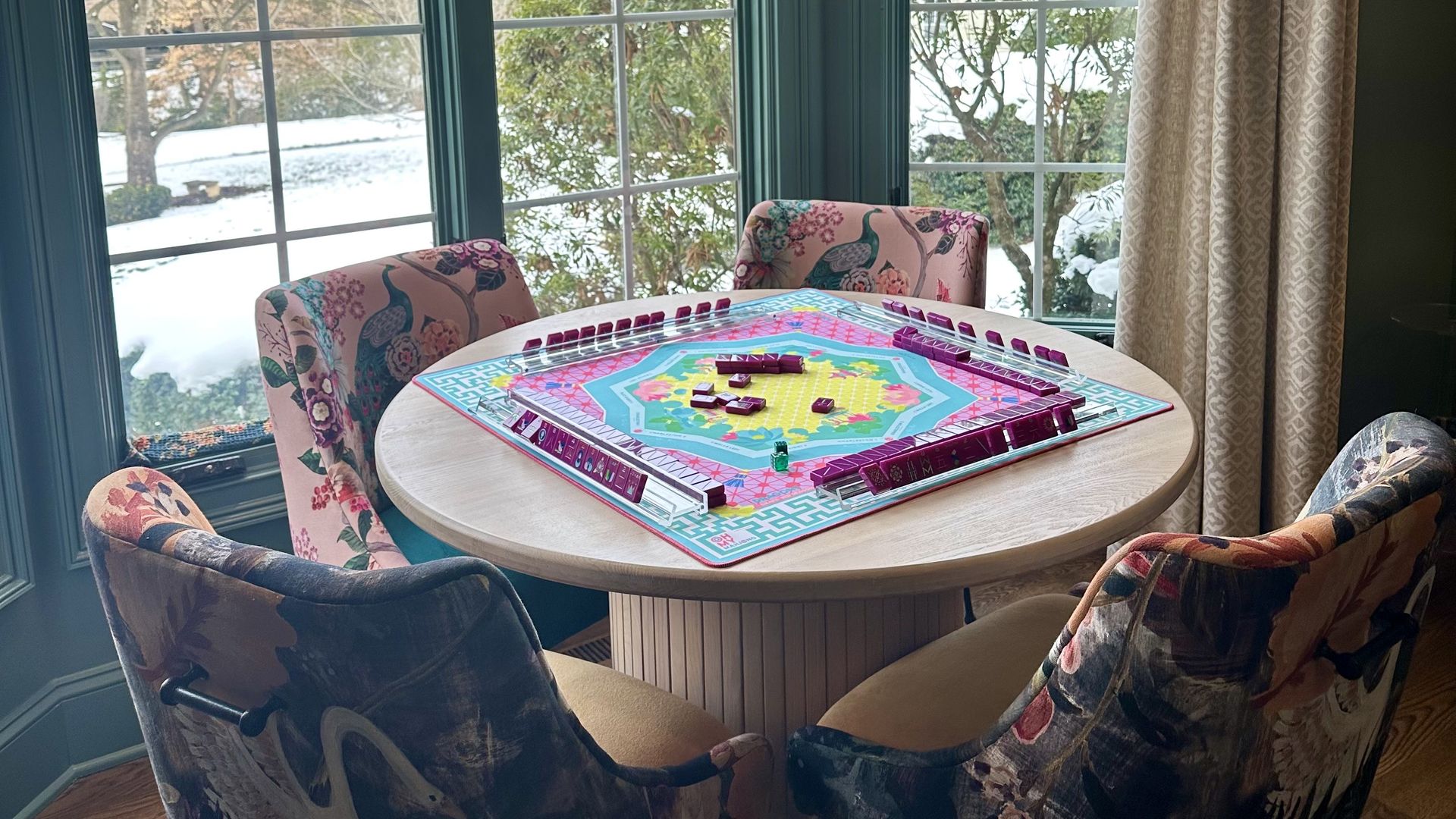A mahjong room in Charlotte. Round wooden table with colorful board game set in a corner room with large blue-framed windows showing a snow-covered yard and green bushes. Four floral upholstered chairs surround the table.