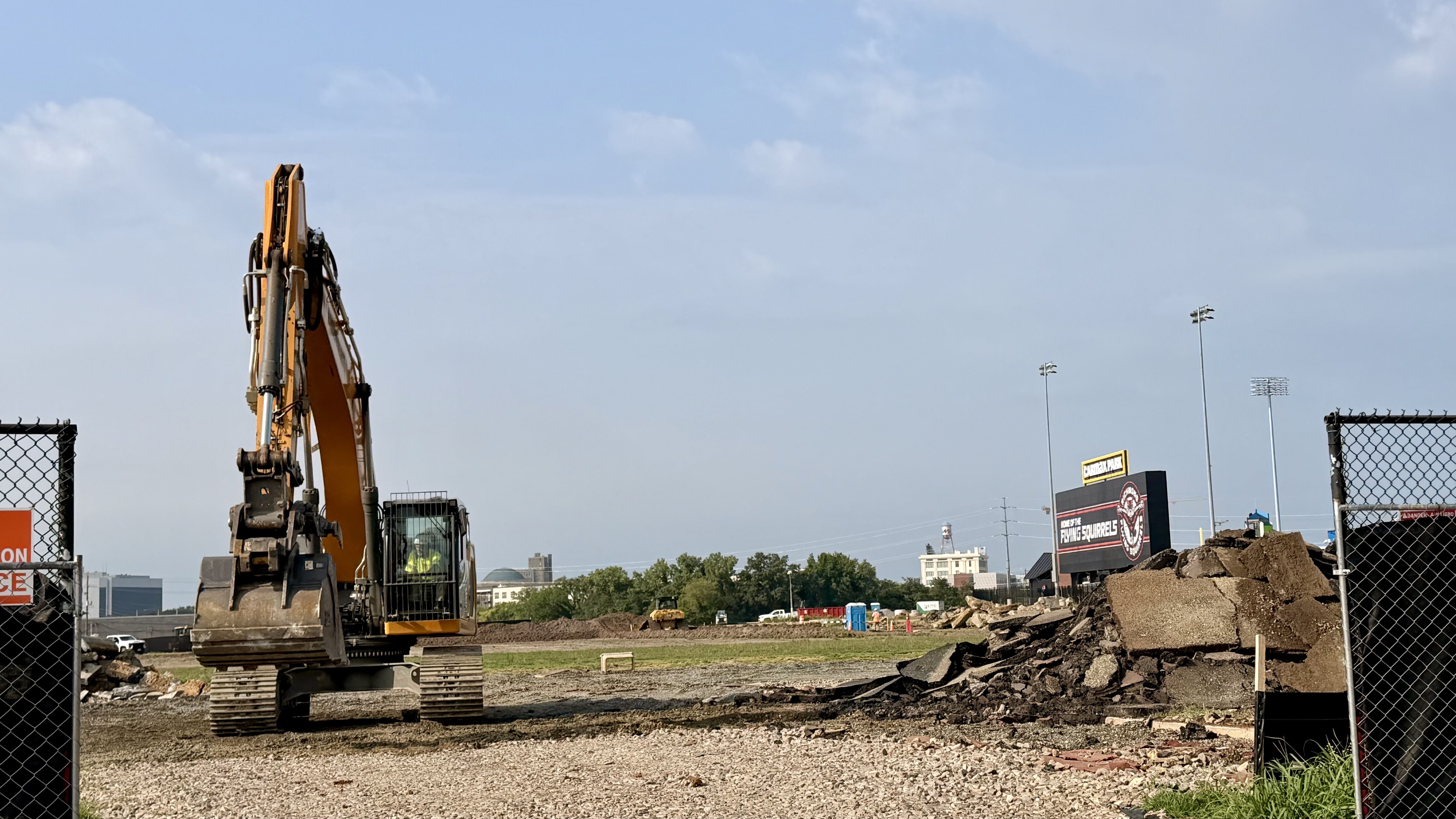Orange construction excavator with extended arm sits on a dirt site behind a chain-link fence; piles of dirt and distant buildings and billboards line the background under a clear blue sky.