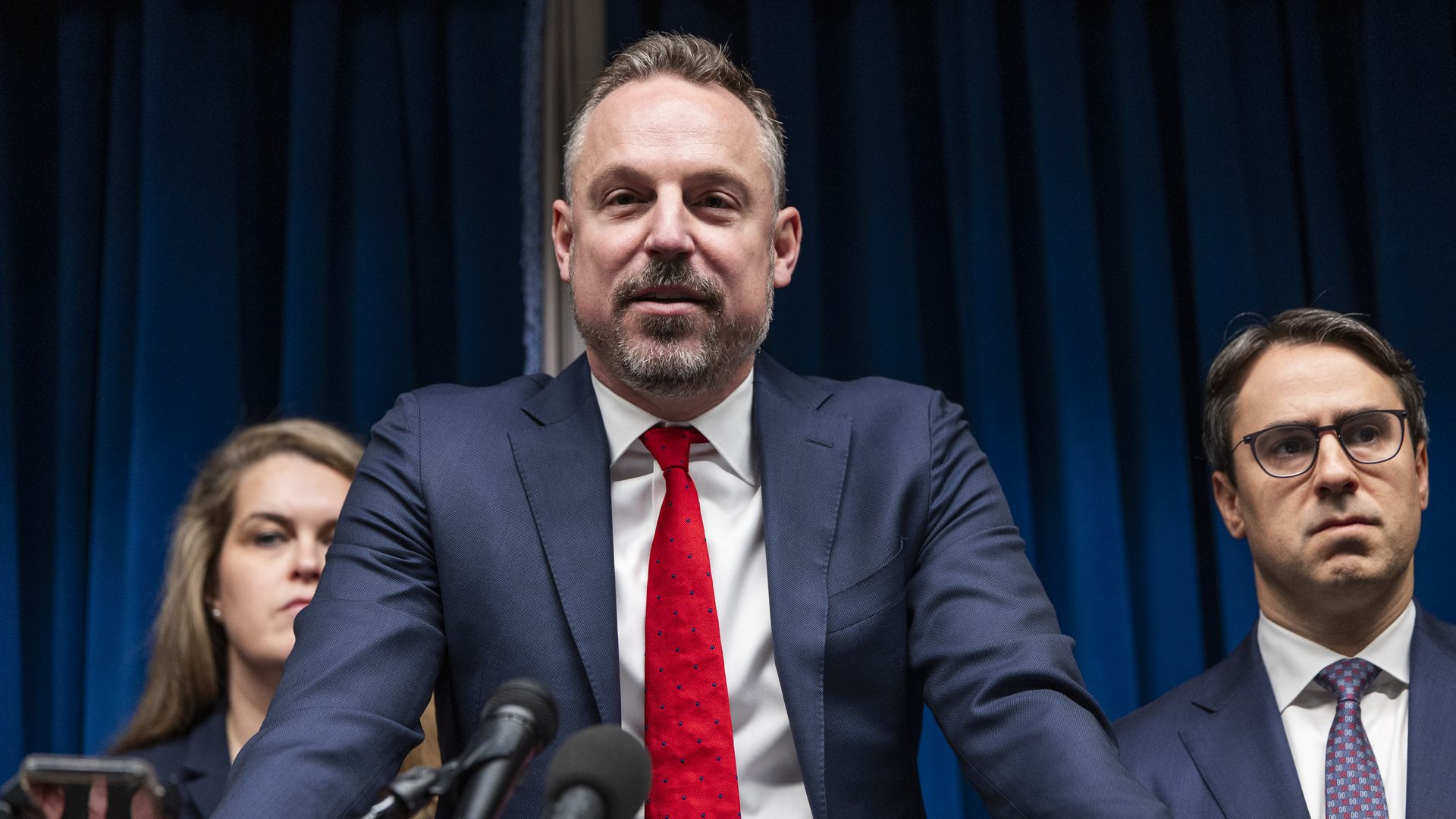 Joe Thompson, wearing a blue suit and red tie, stands at a lectern flanked by other officials. 