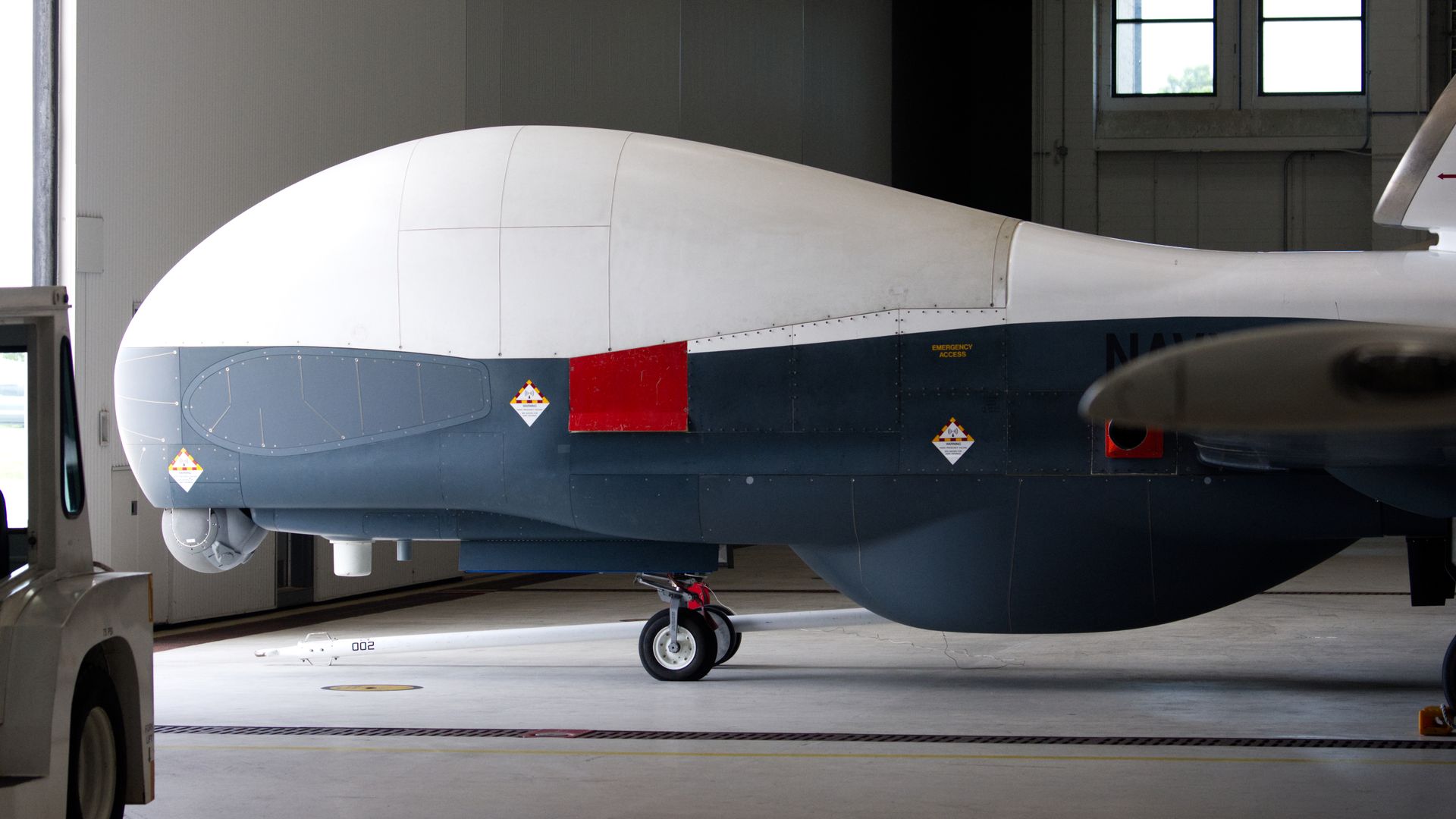 A large white-and-grey drone sits inside a hangar. The building is well lit with bright sunlight streaming through the doors and windows.